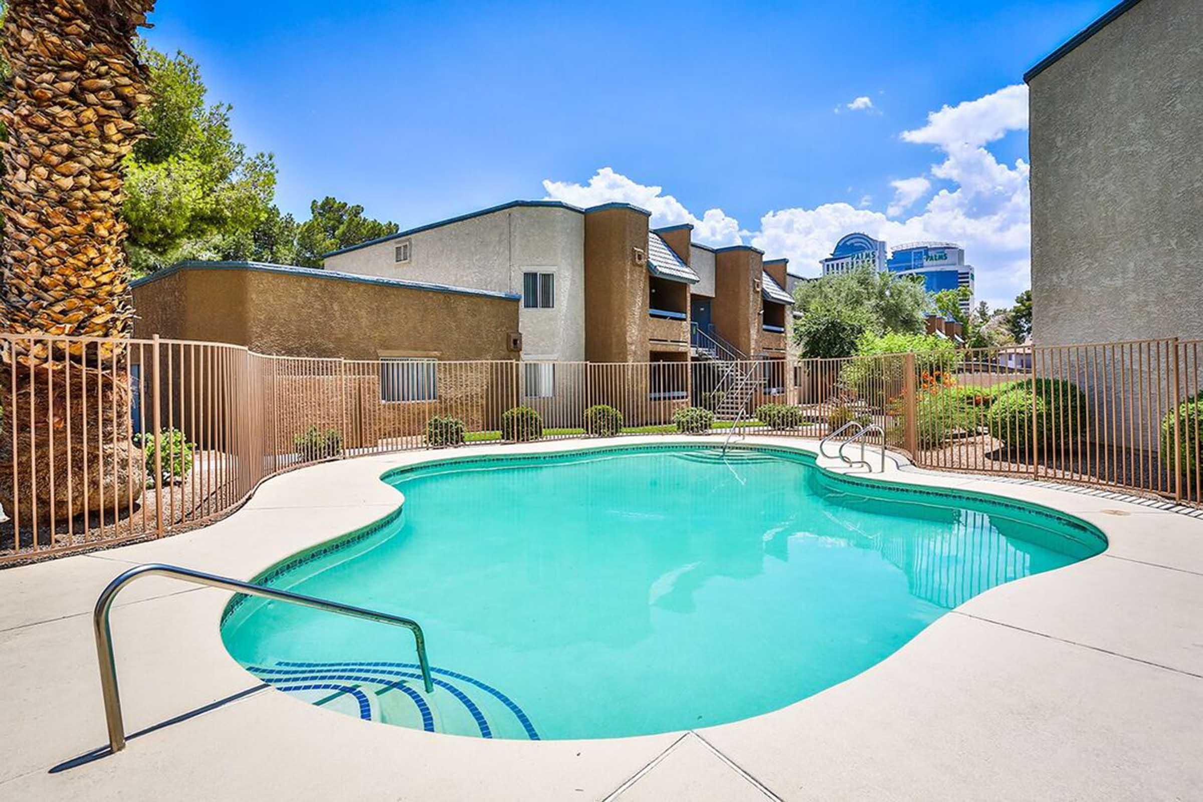 A turquoise swimming pool surrounded by a fence, with landscaped greenery and palm trees. The pool area is set against a backdrop of residential buildings under a clear blue sky with a few clouds. The scene is inviting and tranquil, ideal for relaxation on a sunny day.