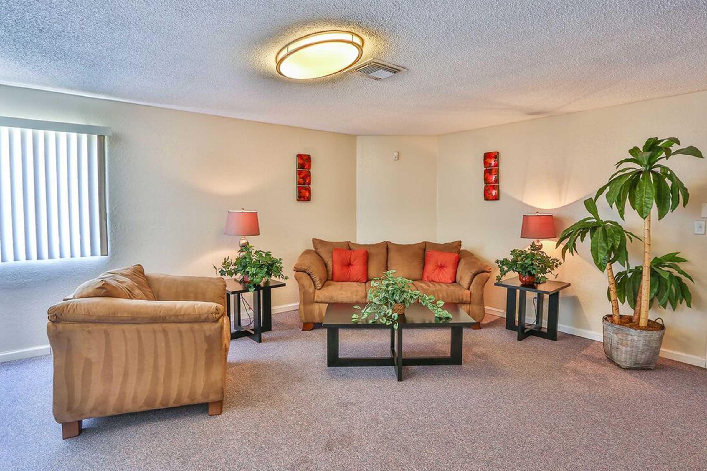 A cozy living room featuring a beige sofa with red cushions, a matching armchair, and a glass coffee table adorned with greenery. Two decorative lamps with shades illuminate the space, while potted plants add a touch of nature. The walls are light-colored, and sunlight filters through vertical blinds.