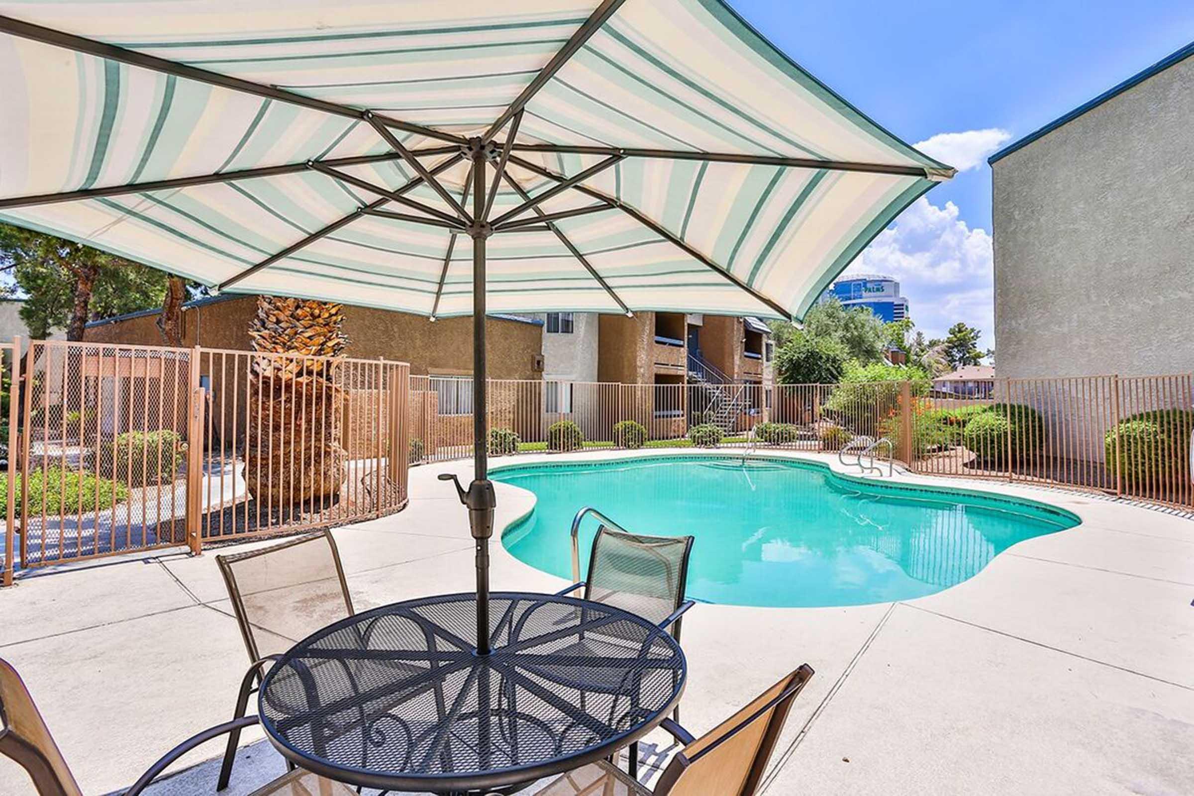 A swimming pool surrounded by a gated area, featuring a patio table with chairs and a large striped umbrella. Lush greenery and a few residential buildings are visible in the background, under a clear blue sky with scattered clouds.