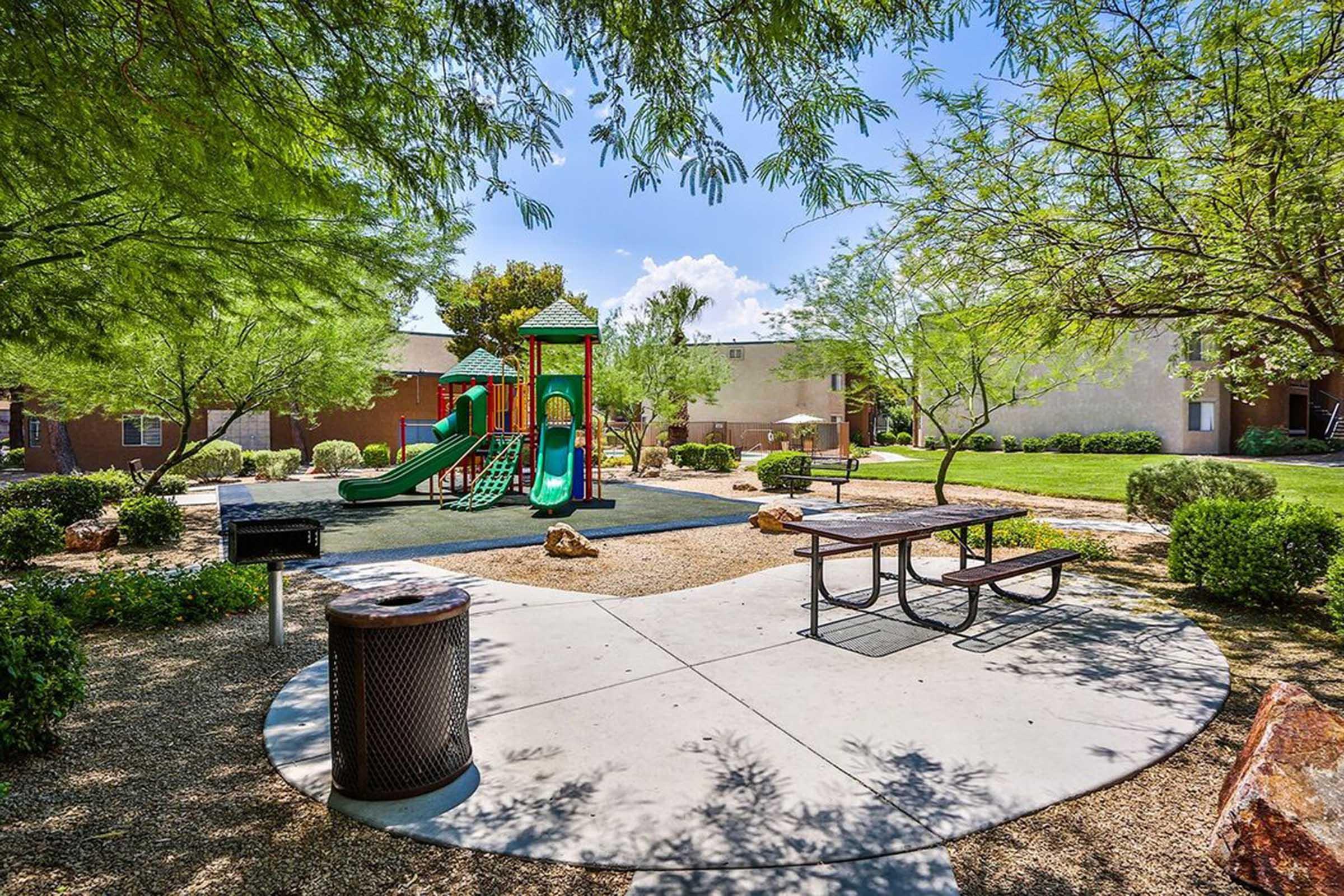 A sunny park scene featuring a colorful playground with slides, surrounded by green trees and shrubs. There is a picnic table in the foreground and a trash can nearby, with a grass area and residential buildings in the background.