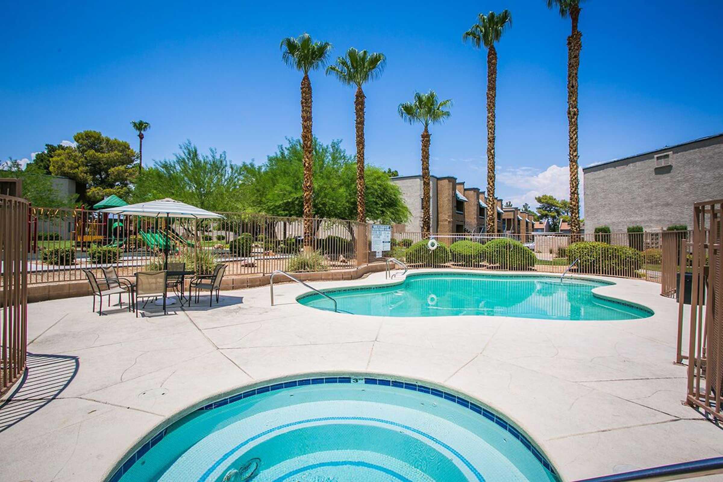 A sunny outdoor pool area surrounded by palm trees and green shrubs. There are lounge chairs and umbrellas set up nearby, with apartment buildings in the background. The sky is clear and blue, creating a relaxing atmosphere.