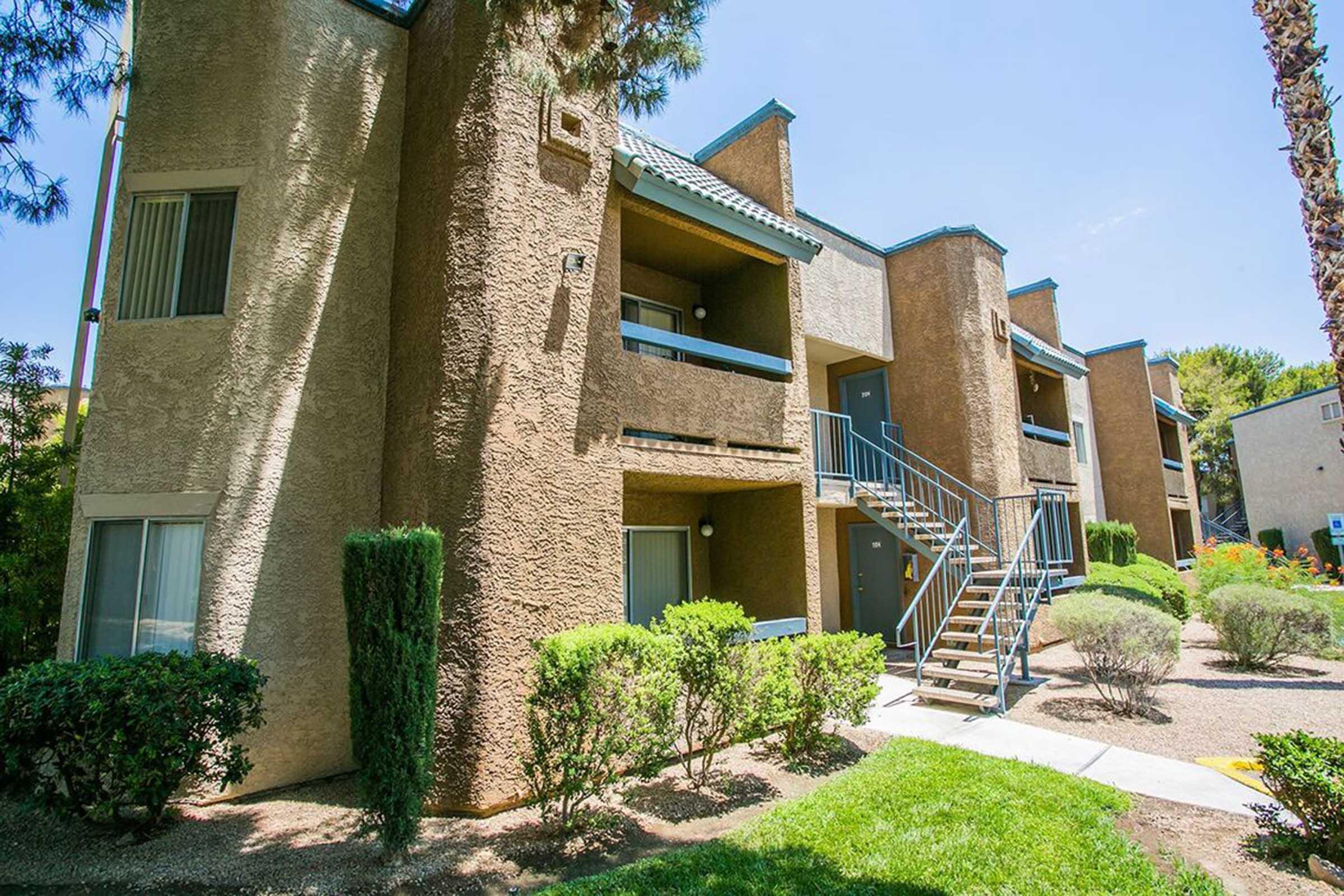 A two-story apartment building with beige and brown exterior walls. The building features balconies, a set of stairs, and landscaping with shrubs and trees. The sky is clear and blue, indicating a sunny day.