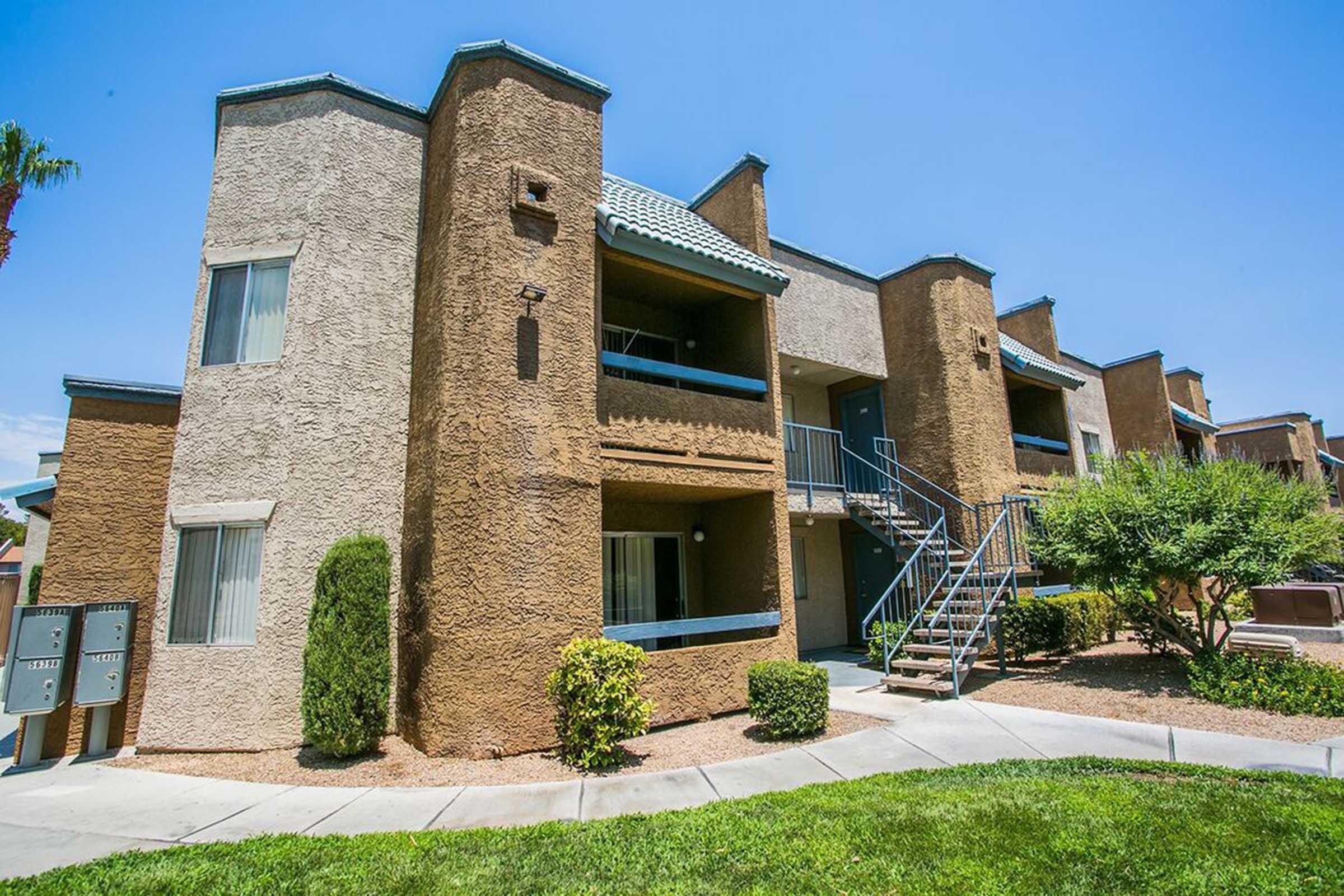 A two-story apartment building with a textured exterior in light brown and beige. The building features balconies with blue railings, surrounded by landscaped greenery and a concrete pathway. There's a set of mailboxes in front, and clear blue skies above.
