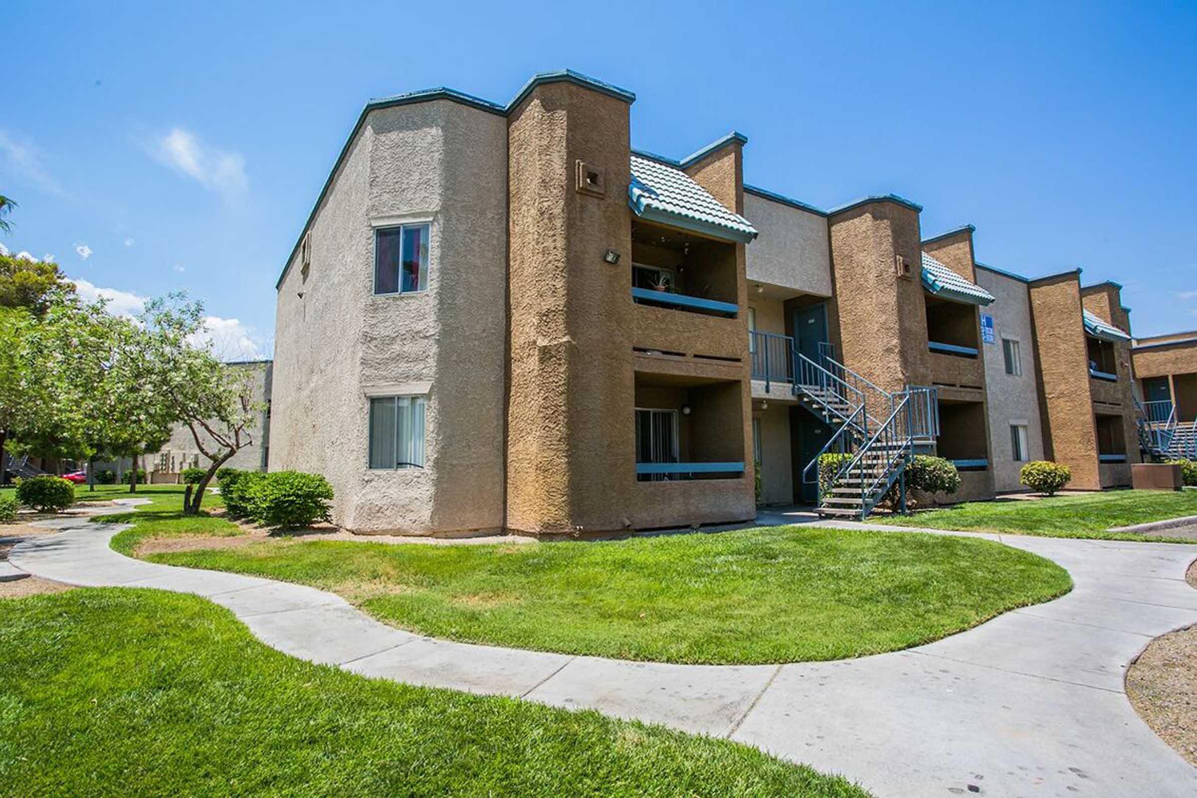 A multi-unit apartment building with a mix of brown and beige exteriors, featuring green grass, landscaped pathways, and blue skies in the background. The building has several balconies and is surrounded by small trees and shrubs, providing a serene residential environment.
