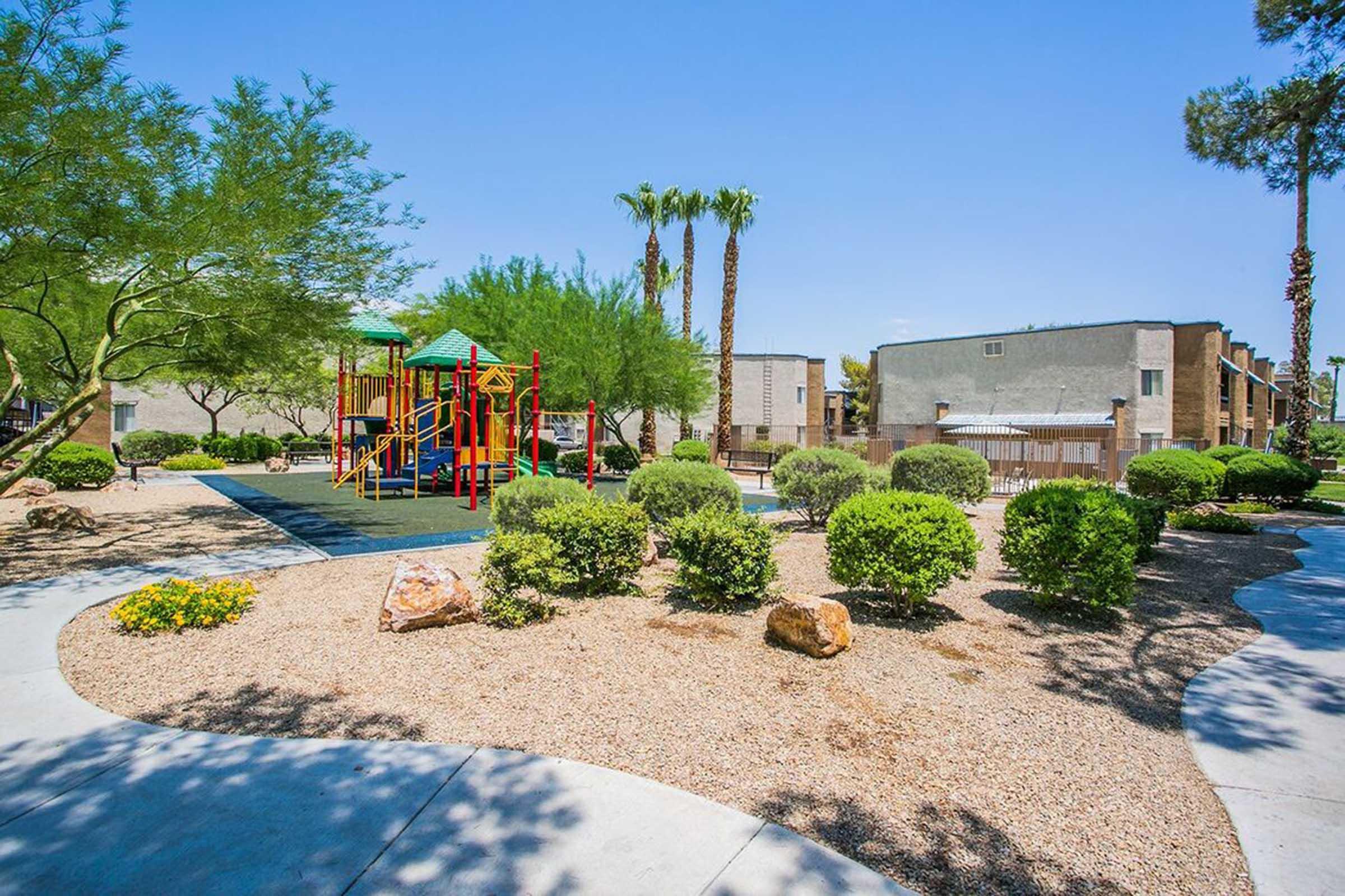 A sunny playground area featuring colorful equipment surrounded by green shrubs and trees. A winding path leads through a landscaped area with rocks and flowers, providing a pleasant outdoor environment. Apartment buildings are visible in the background under a clear blue sky.