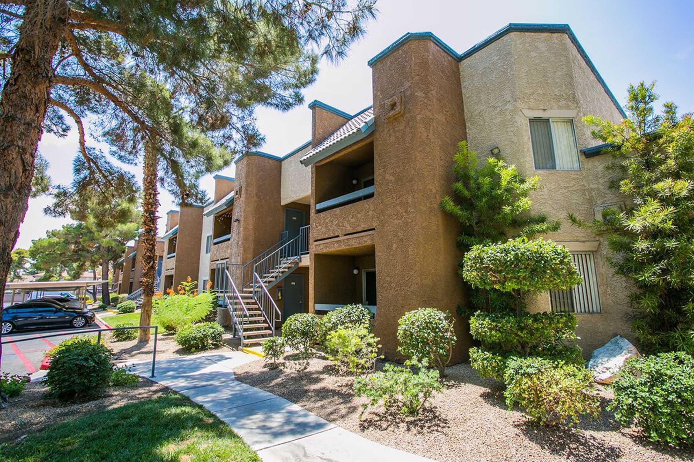 A view of a modern apartment complex featuring three-story buildings with balconies. Lush greenery and neatly maintained shrubs surround the area, while a stone pathway leads through the landscaping. A parking lot with a car is visible in the foreground, under clear blue skies.