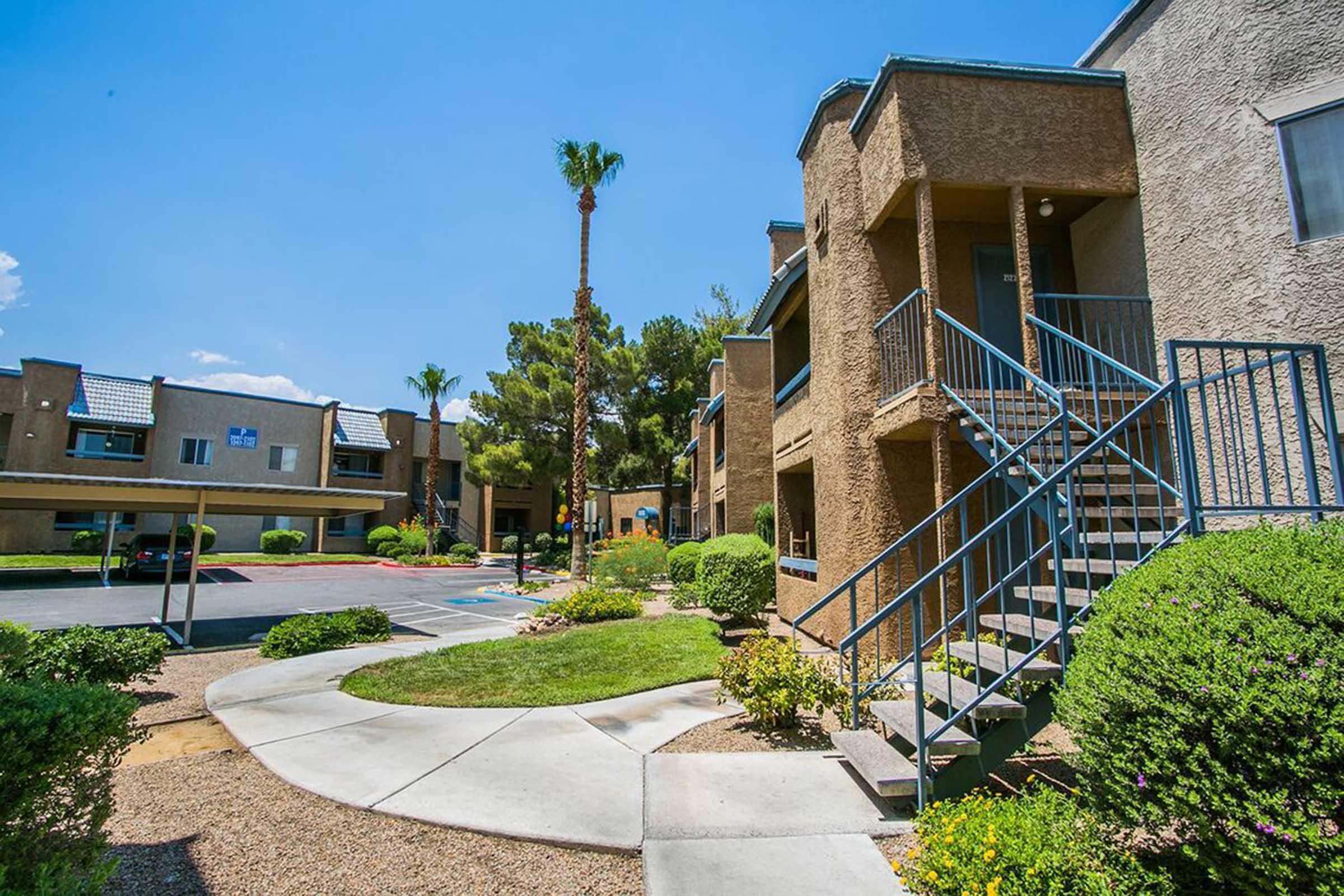 A landscaped area of a residential complex featuring palm trees, shrubs, and flower beds. The buildings are two stories with staircases leading to second-floor entrances. Clear blue sky above and a parking lot in the background.