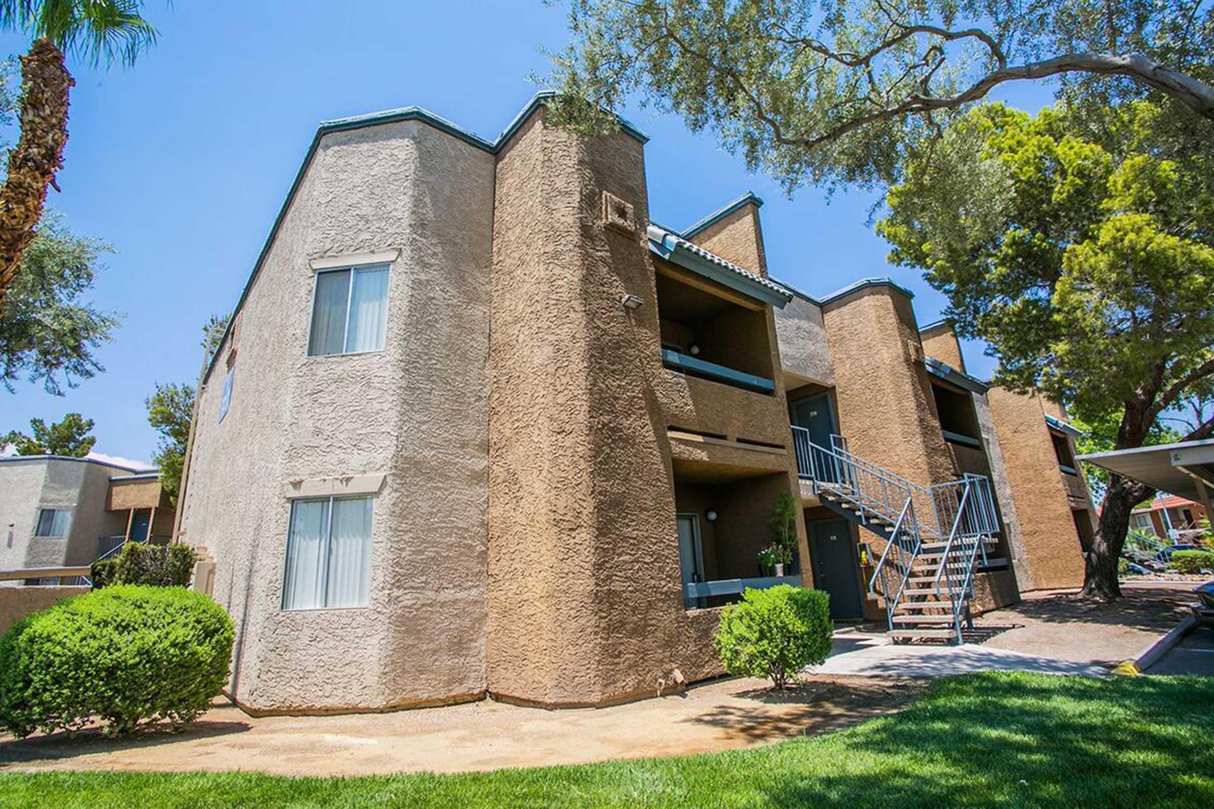 A two-story apartment building with a rough textured exterior in a residential area. The building features balconies, steps leading to the upper floor, and is surrounded by well-maintained greenery, including small bushes and trees, under a clear blue sky.