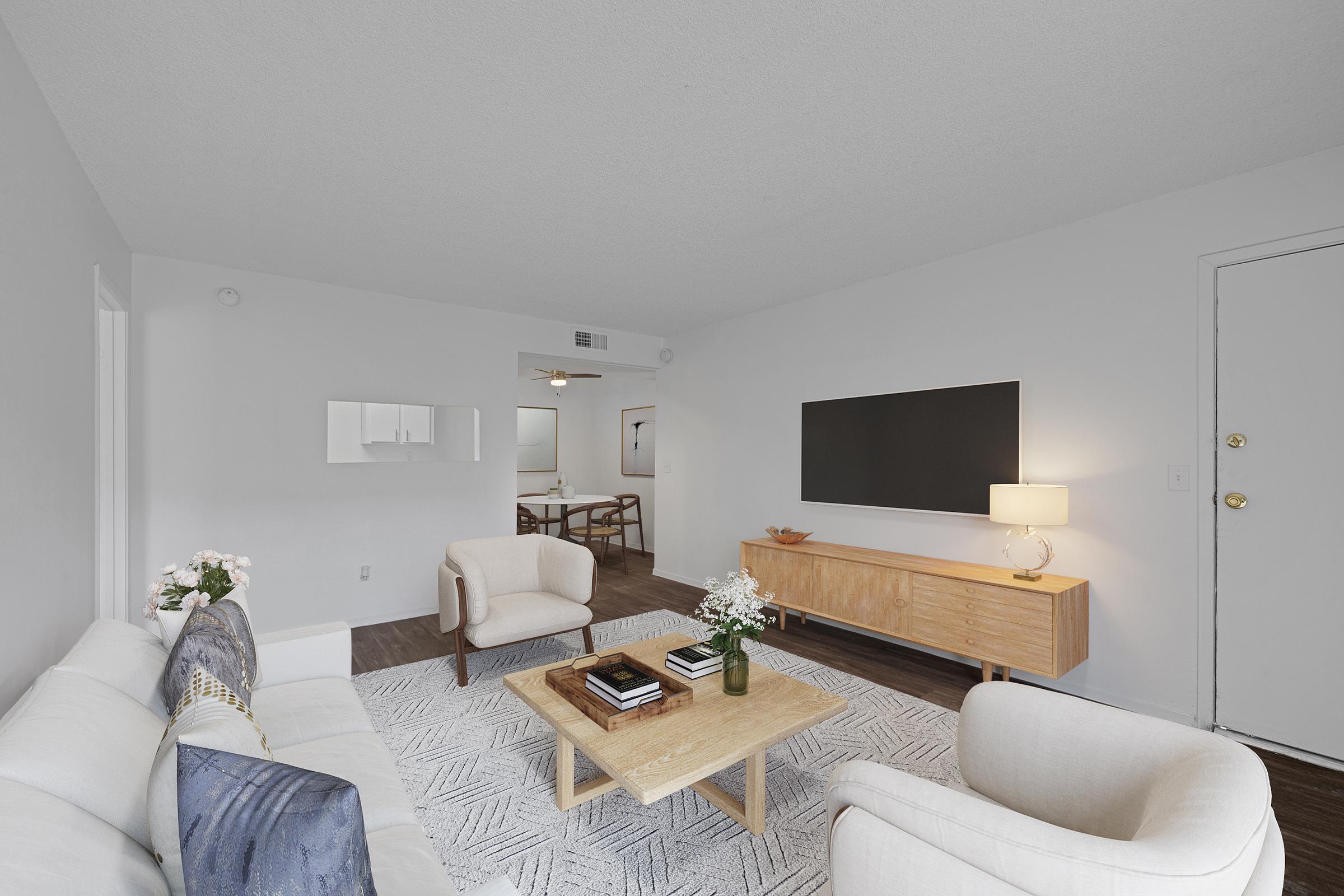 A modern living room featuring a light-colored sofa with decorative pillows, a wooden coffee table with magazines, and a side table with a lamp. A mounted flat-screen TV is on the wall, and there is a textured area rug on the floor. The space is bright with minimal decor, creating a clean and inviting atmosphere.