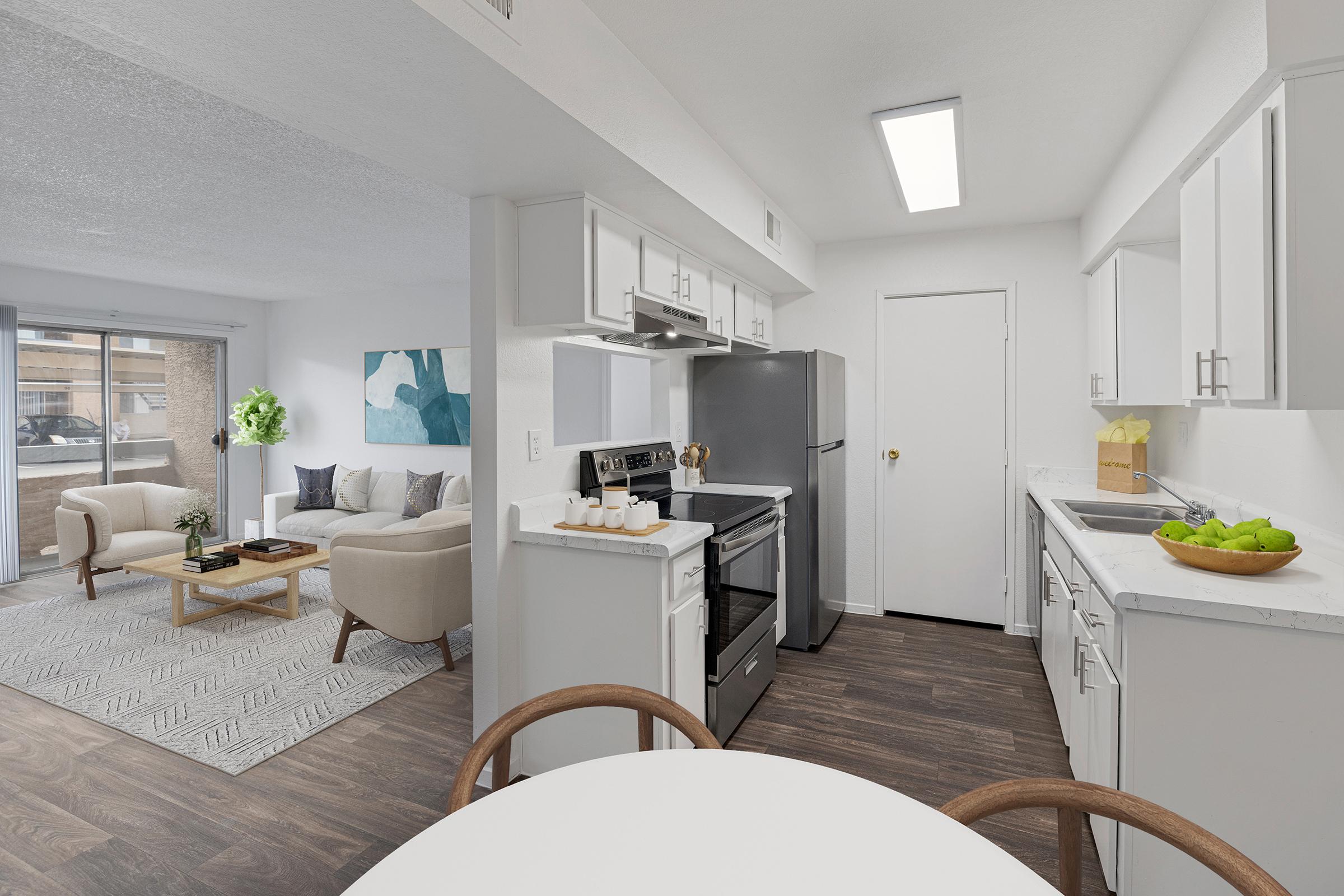 Modern kitchen and living room in an open-concept apartment. The kitchen features stainless steel appliances, white cabinets, and a countertop with green apples. The living area has a cozy sofa, coffee table, and a large window allowing natural light. A decorative blue and white painting is on the wall.