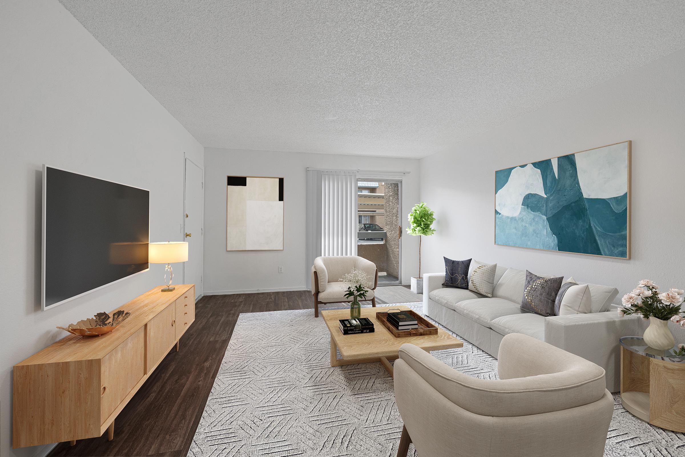A modern living room featuring a light color palette. The space includes a white sofa with decorative pillows, two armchairs, a wooden coffee table, and a sideboard with a lamp. A flat-screen TV is mounted on the wall, and large windows bring in natural light. Artwork adorns the walls, enhancing the contemporary decor.