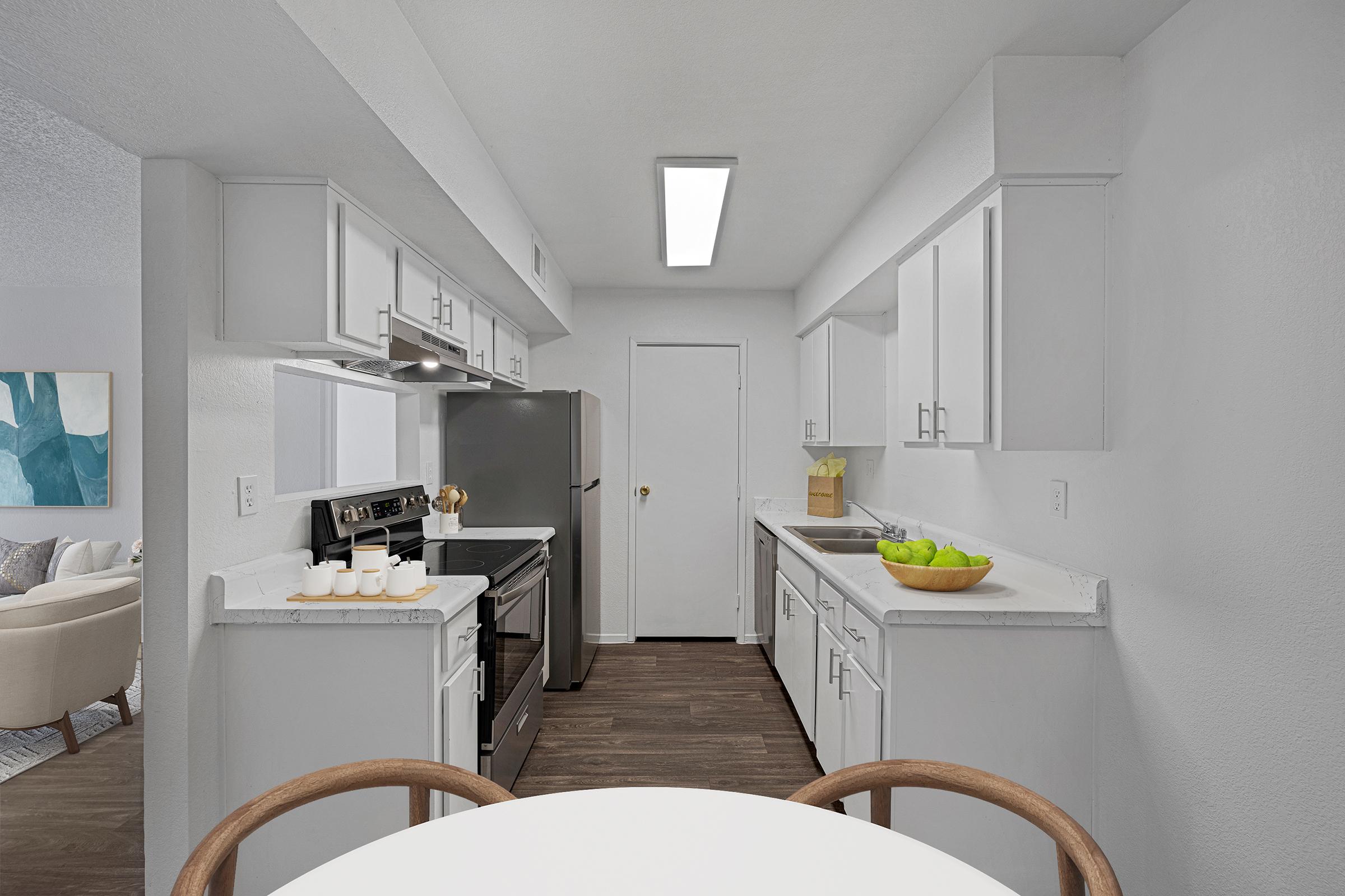 A modern kitchen with white cabinets, stainless steel appliances, and a light countertop. A round table with wooden chairs is positioned in the foreground, while a bowl of green fruits is on the table. Natural light enters through a skylight, creating a bright and airy atmosphere.