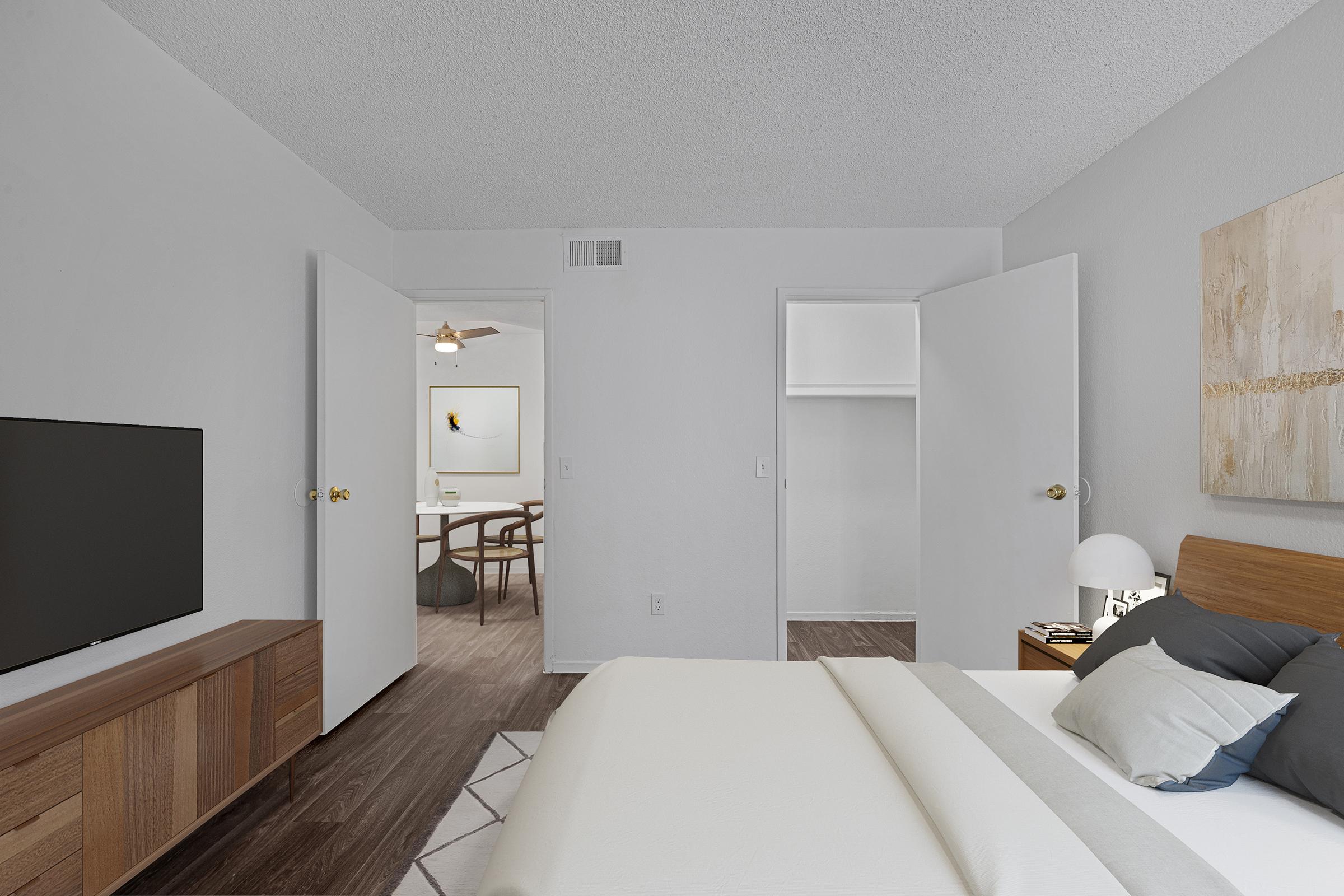 A cozy bedroom featuring a wooden bed with light bedding, a nightstand, and a lamp. A flat-screen TV is mounted on a wooden dresser. The room has two open doors leading to adjacent spaces, with a simple, neutral color scheme and modern decor elements. Bright natural light fills the space.
