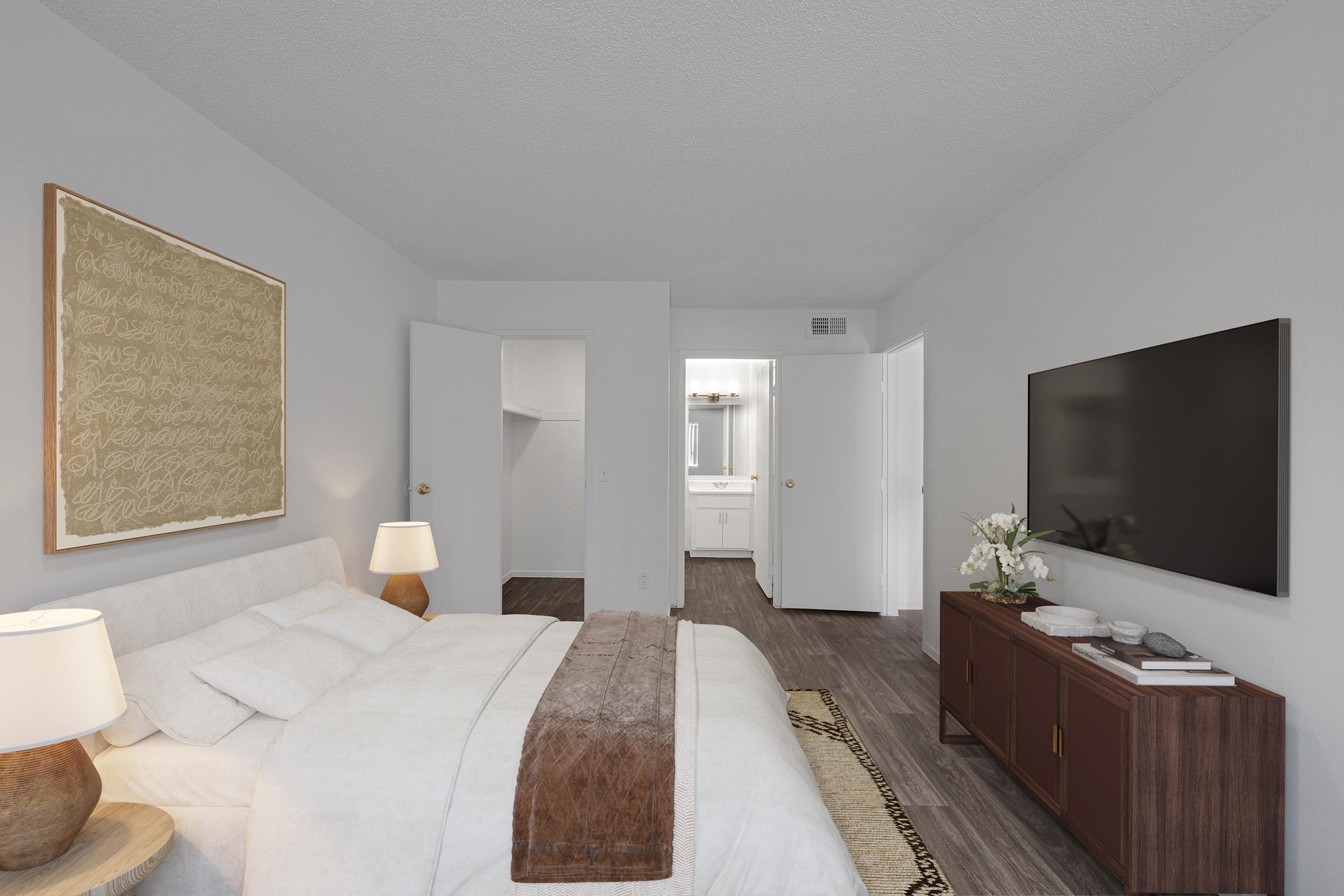 A modern bedroom featuring a bed with white linens and a brown throw. There are two bedside lamps, a large TV mounted on the wall, and a wooden dresser. The room has neutral-colored walls and hardwood flooring, with an open doorway leading to a bathroom area visible in the background.