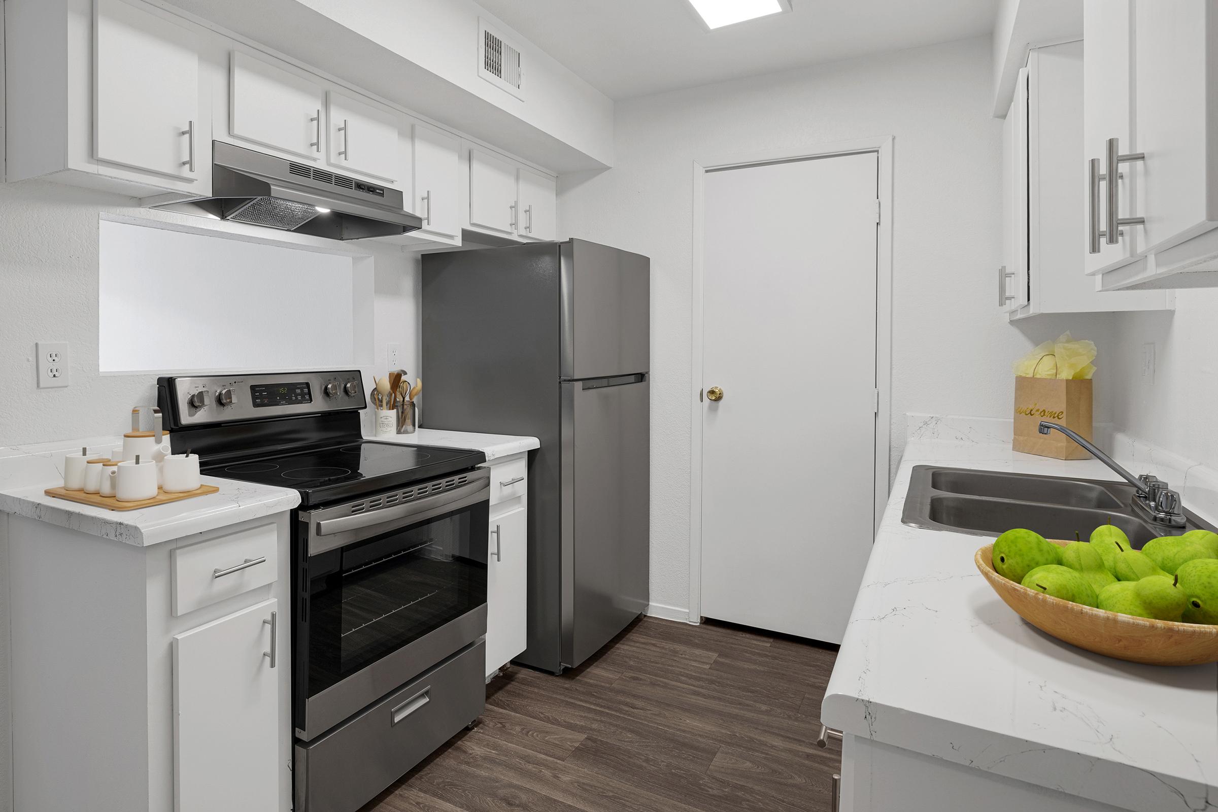 A modern kitchen featuring white cabinets, stainless steel appliances, and a countertop with a bowl of green apples. There is an oven and a stove, a refrigerator, and a sink next to a door leading to another room. The overall design is clean and minimalistic.