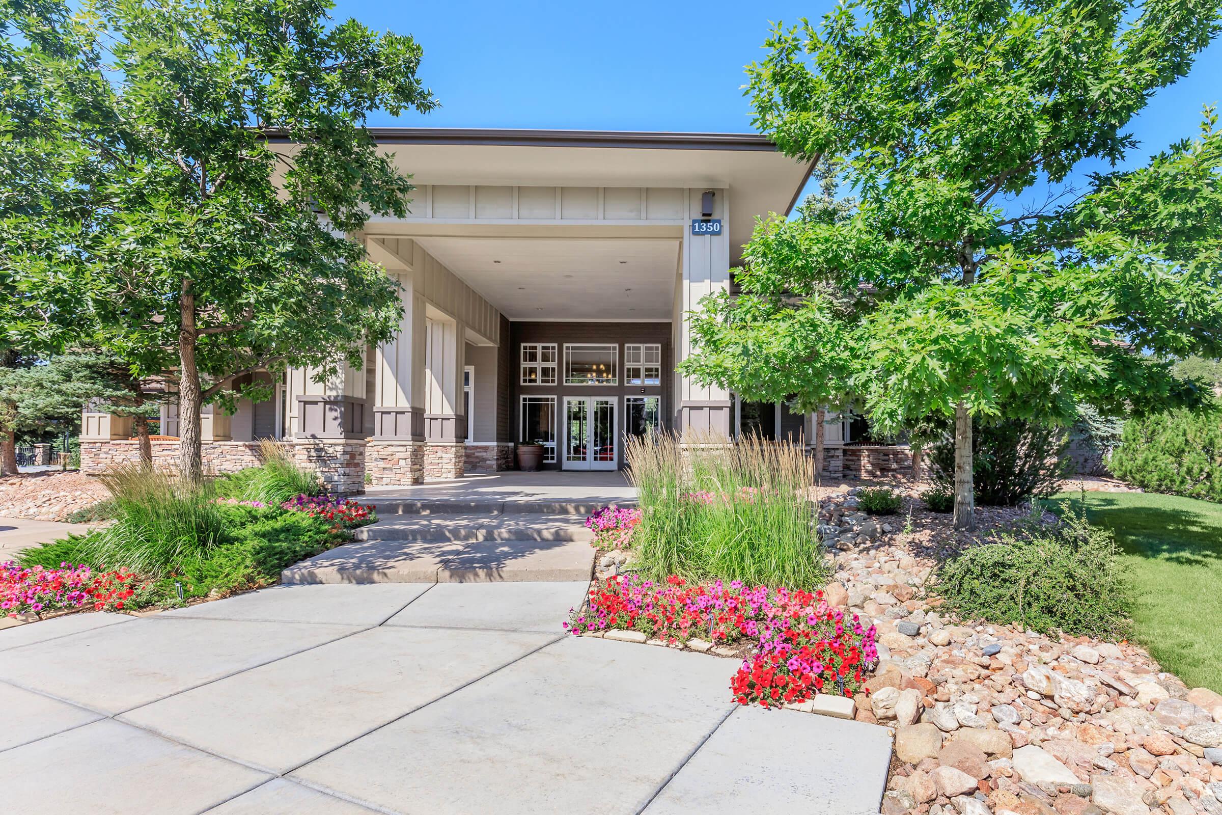 A modern building entrance surrounded by lush greenery and colorful flowerbeds. The pathway leads up to the doors, flanked by trees and shrubbery, all under a clear blue sky. The architecture features a combination of stone and wood elements, creating an inviting atmosphere.
