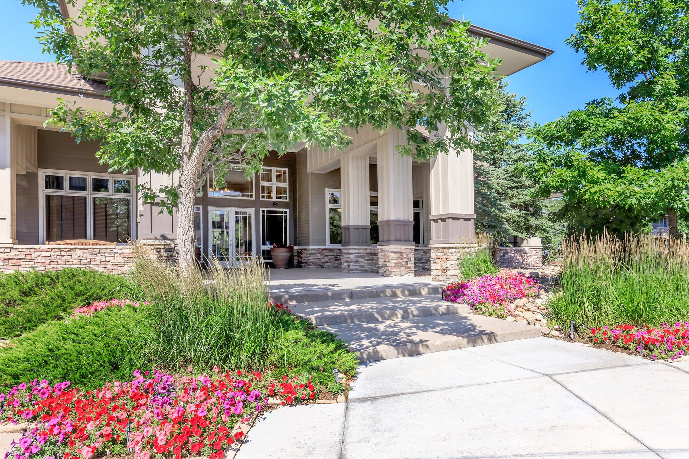 A sunny view of a modern building entrance surrounded by vibrant flowers, green shrubs, and trees. The pathway leads to the entrance, which features large windows and a welcoming facade. The landscape design includes colorful blooms and well-maintained greenery, creating an inviting atmosphere.