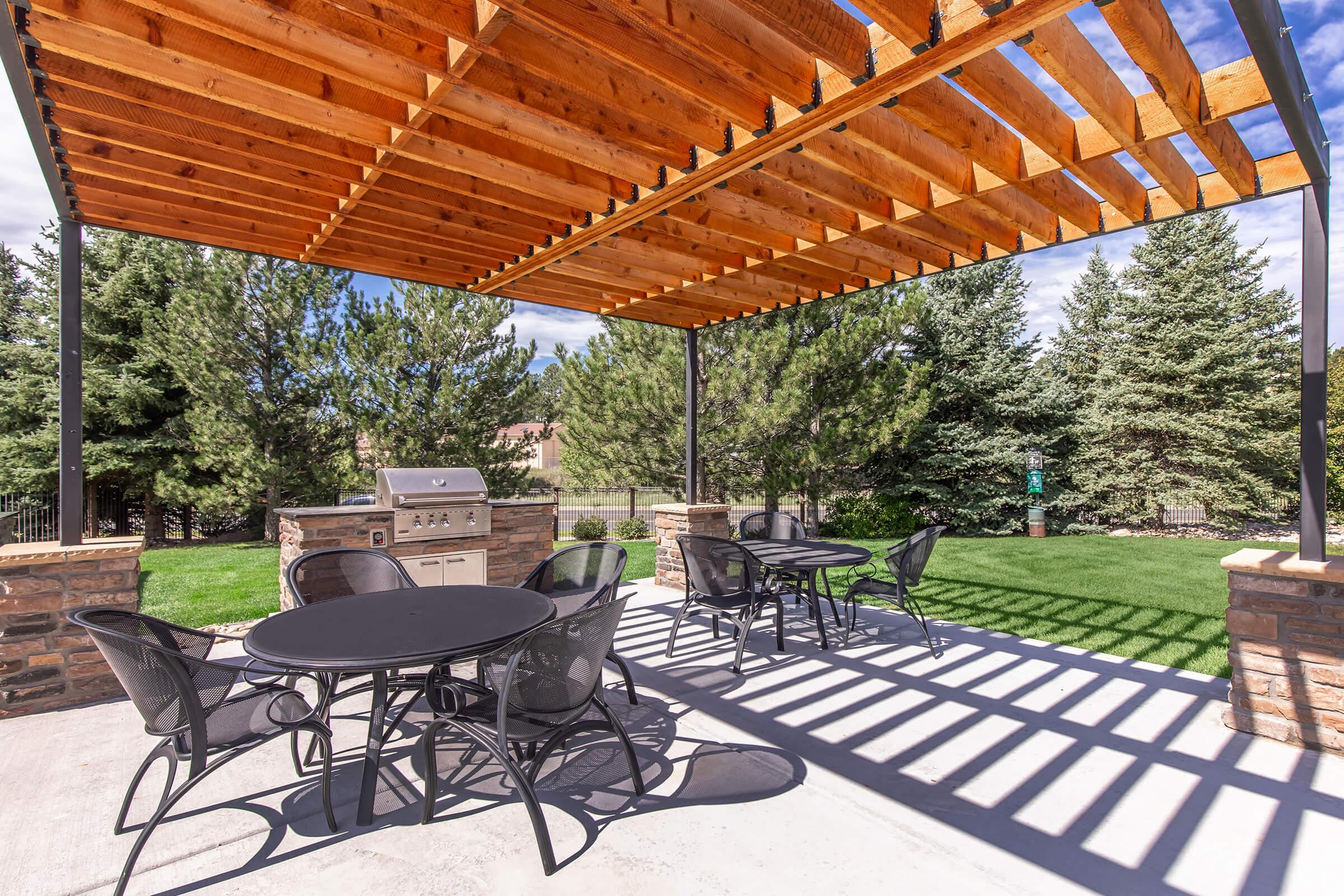 Outdoor patio area featuring a wooden pergola overhead, a stone grill station, and several black metal chairs and tables arranged on a concrete surface. Lush green grass and mature trees provide a scenic backdrop.