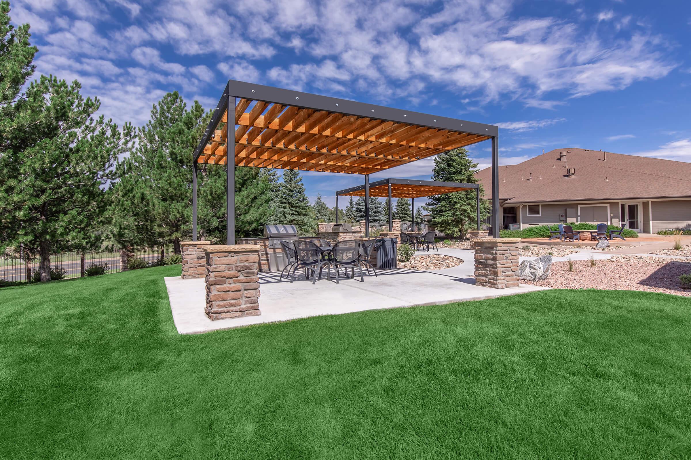 A spacious outdoor patio featuring a pergola with wooden beams, surrounded by well-manicured green grass, trees in the background, and a residential home nearby under a vibrant blue sky with fluffy clouds.