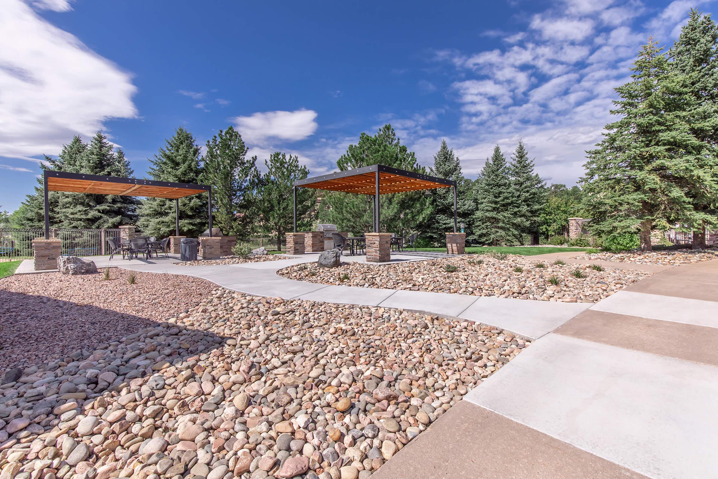 A landscaped outdoor area featuring two covered pavilions with wooden roofs, surrounded by tall green trees. The ground is covered with a path of concrete and decorative rocks, creating a serene environment under a bright blue sky with scattered clouds.