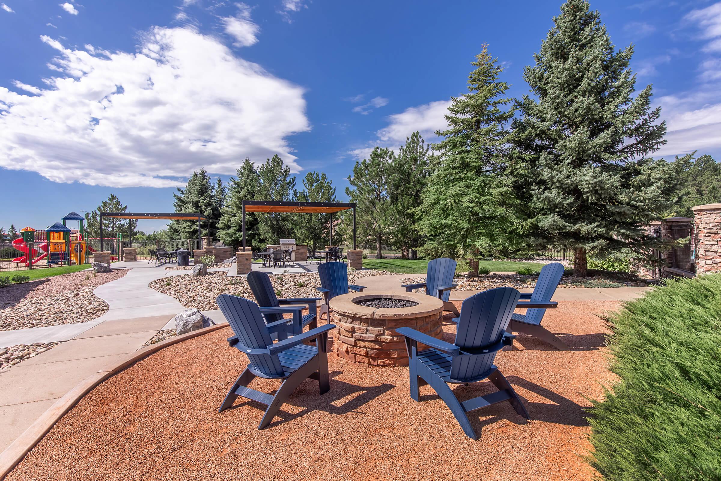 A landscaped outdoor area featuring a circular stone fire pit surrounded by several blue Adirondack chairs. In the background, there are trees, a pathway, and a playground, all under a bright blue sky with clouds.