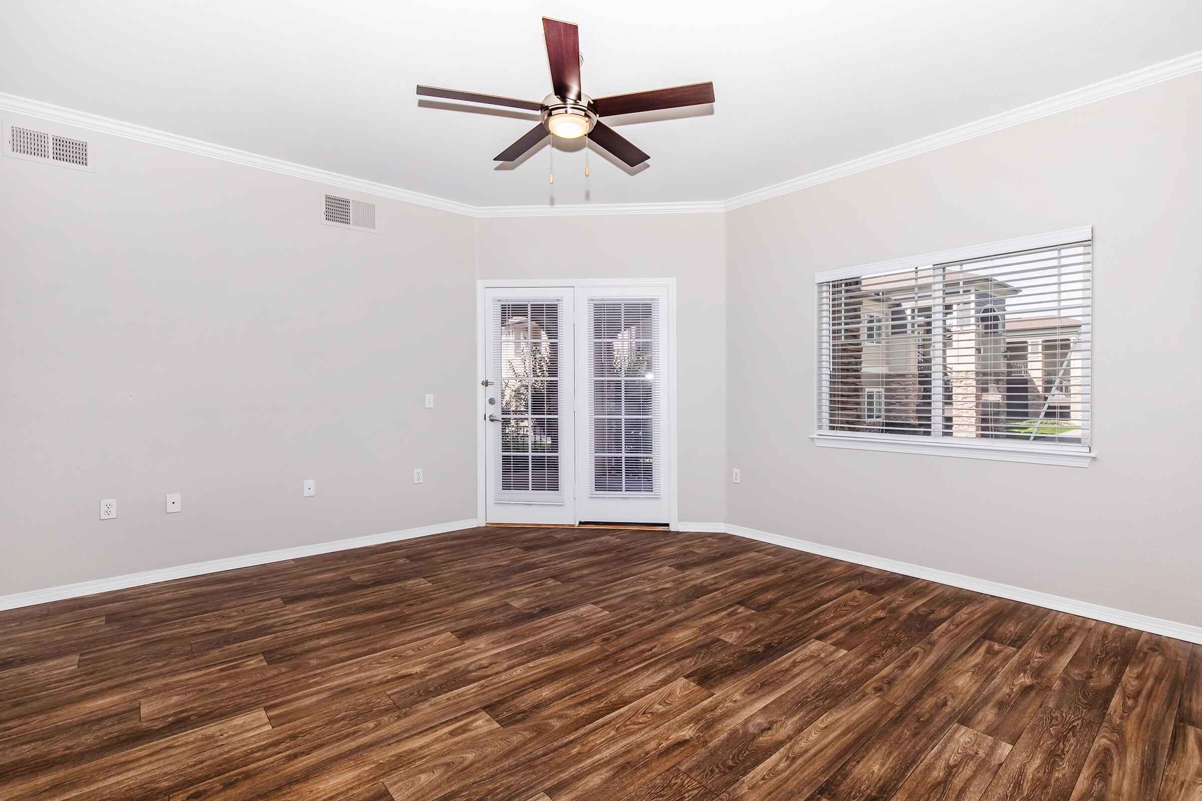 A spacious, empty living room featuring light beige walls, a ceiling fan with wooden blades, and laminate flooring. There are double doors leading outside and a window with horizontal blinds, allowing natural light to fill the room.