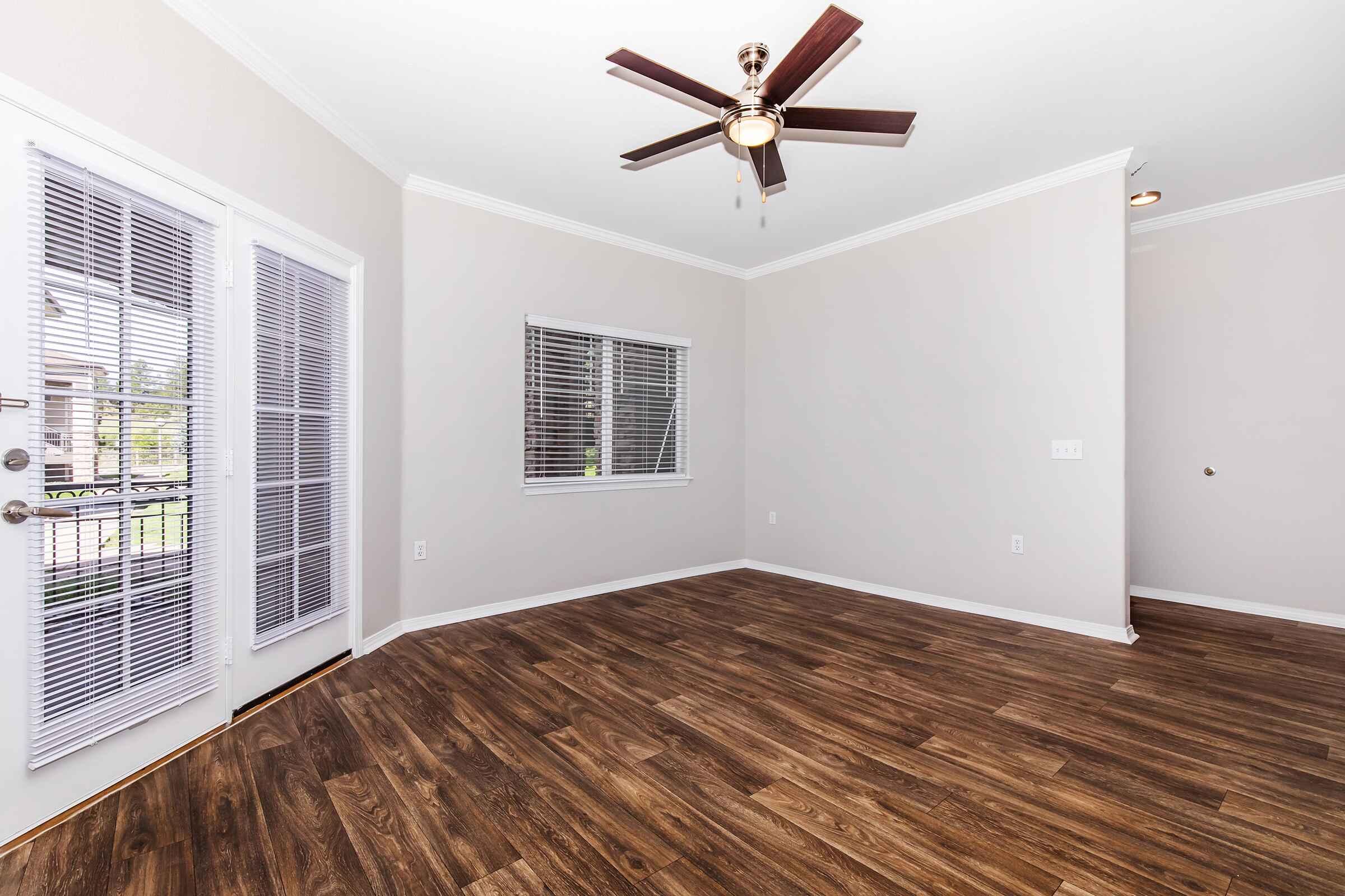 A spacious, empty room with light-colored walls, a ceiling fan, and hardwood flooring. Two pairs of French doors with blinds lead to the outside, and a window provides natural light. The room is well-lit and features a simple, modern design.