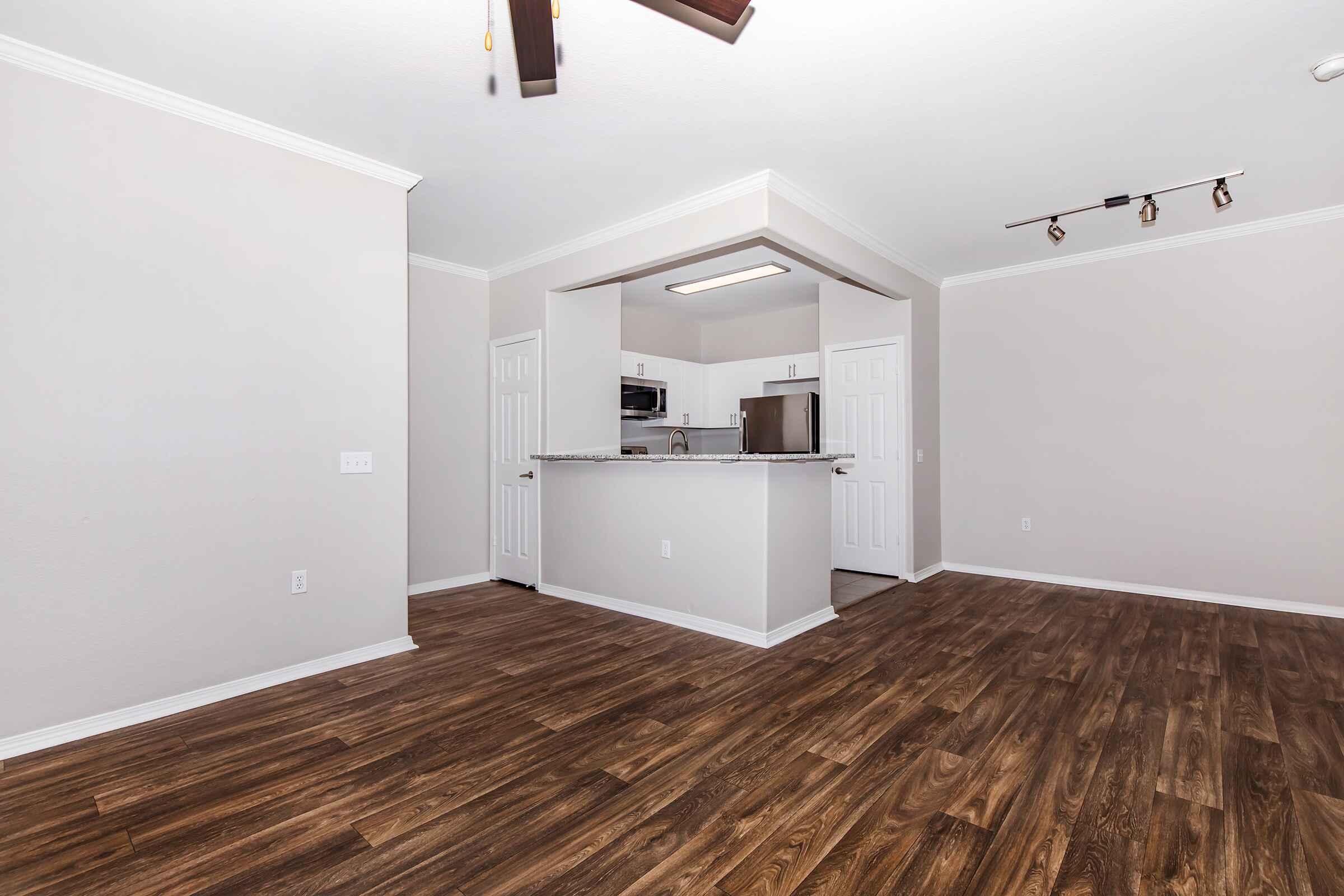 A spacious living area with light-colored walls and dark hardwood flooring. There is an open kitchen with a countertop that faces the living space, stainless steel appliances, and recessed lighting. The room is bright with natural light and features a ceiling fan.