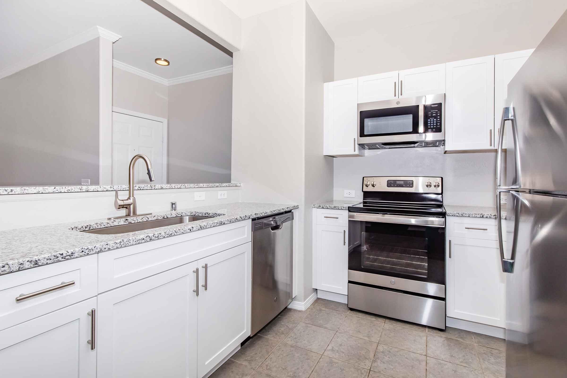 Modern kitchen featuring white cabinetry, granite countertops, and stainless steel appliances, including a microwave, oven, and dishwasher. The space has a clean, contemporary design with neutral walls and tiled flooring, creating an inviting atmosphere.