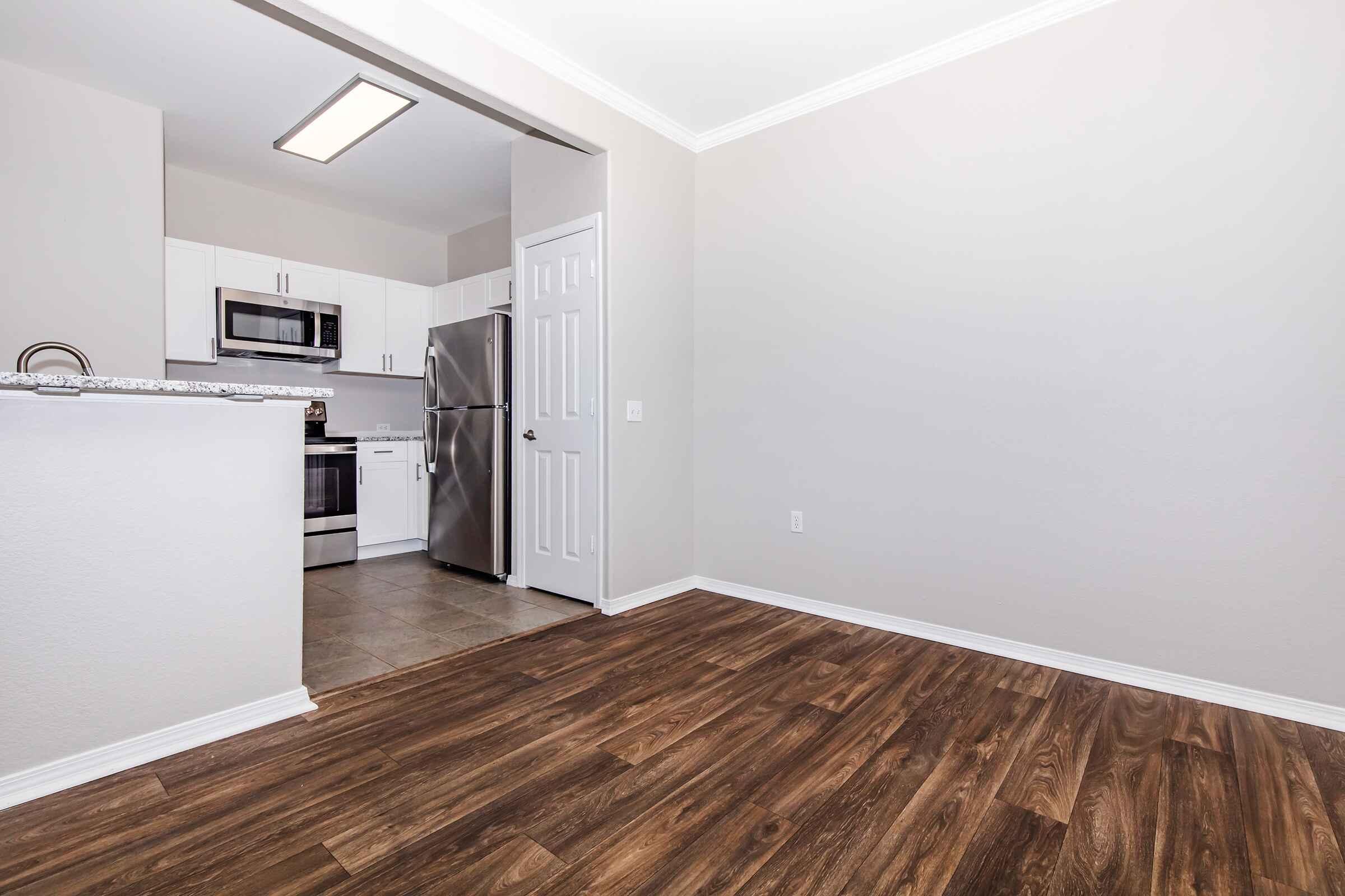 A well-lit room with new hardwood flooring, featuring a kitchen area with white cabinets, stainless steel appliances, and a countertop. The open wall leads to a doorway with a closed door visible. The walls are painted in a light color, creating a modern and inviting atmosphere.