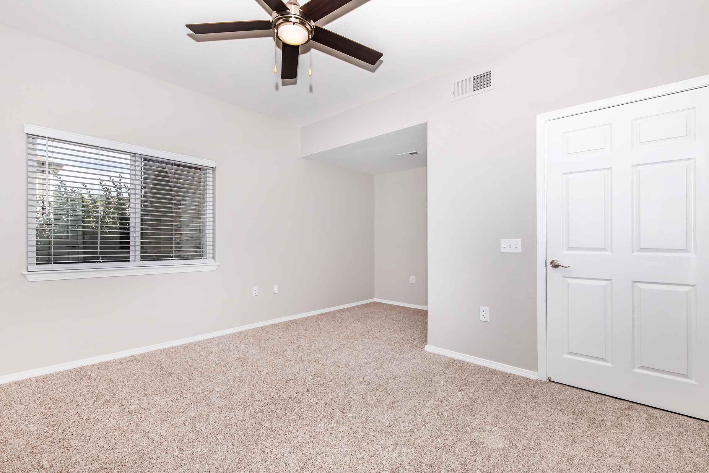 A well-lit, empty room with light-colored walls, carpeted floor, and a ceiling fan. One wall features a window with horizontal blinds. A white door is visible, leading to another space. The room is spacious and clean, offering a neutral setting suitable for various uses.