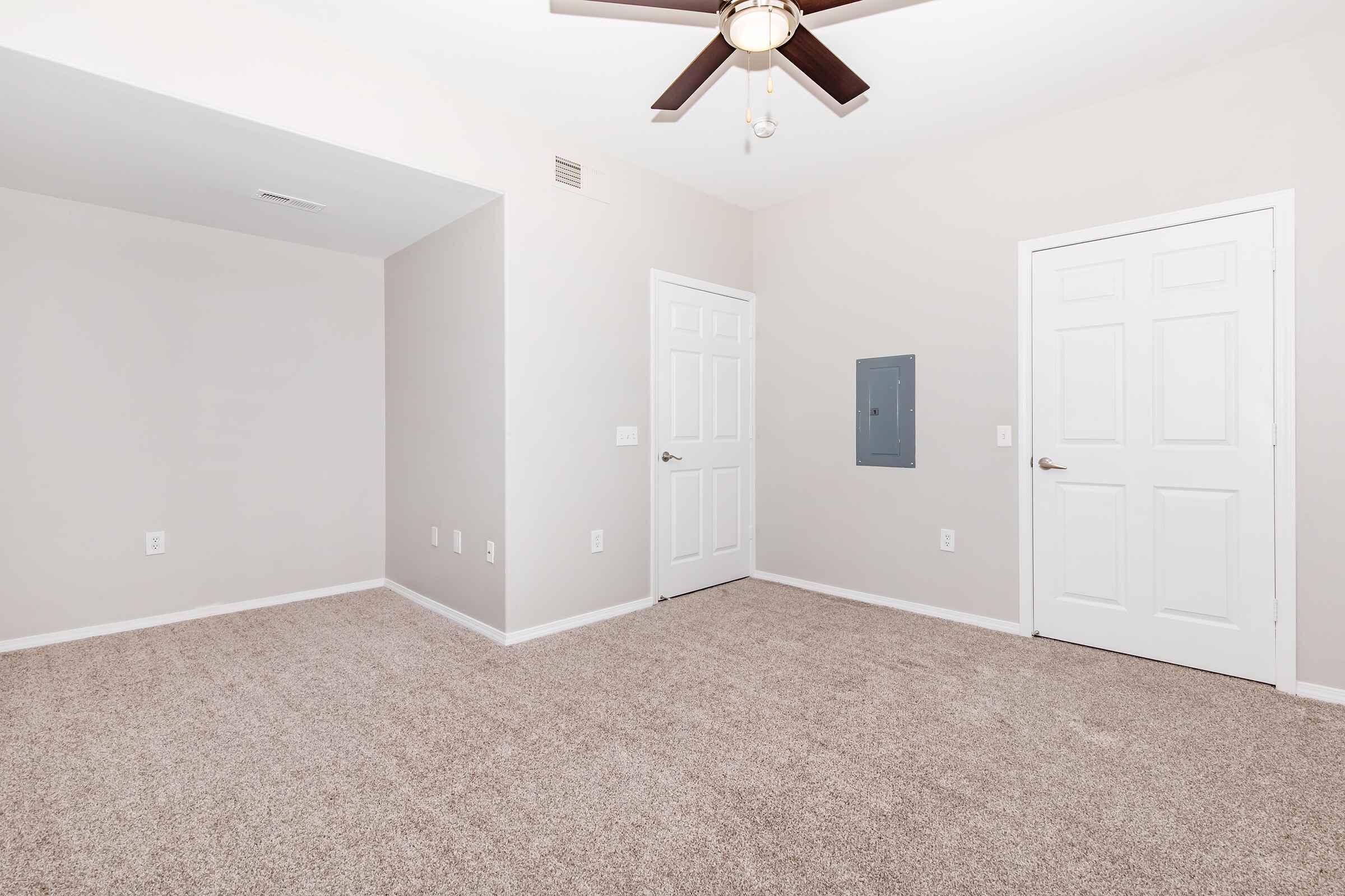 A spacious, empty room featuring light beige walls, carpeted flooring, and a ceiling fan with light. There are two white doors on the right, one leading to a closet and the other likely a standard door. An electrical panel is mounted on the wall next to the doors, and there’s a small nook on the left side.