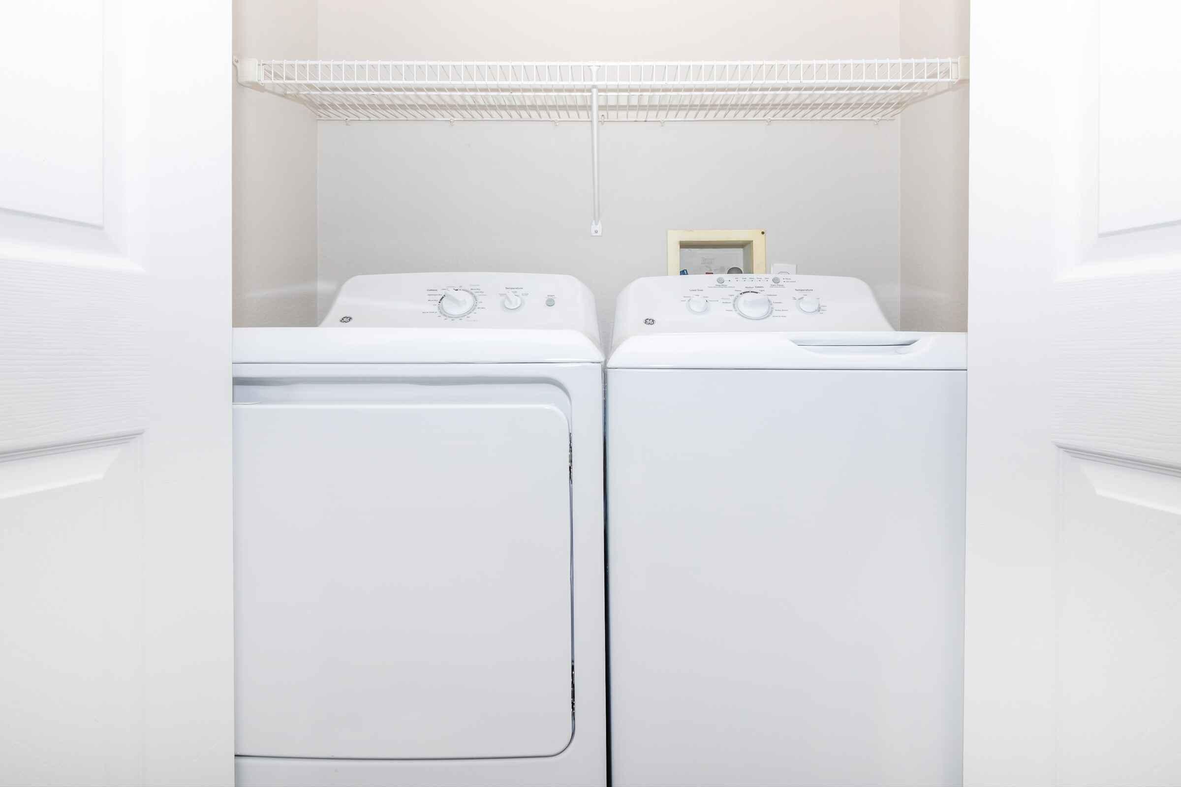 A white washing machine and dryer side by side in a laundry closet, with a shelf above them for storage. The machines feature simple knob controls and a small control panel on the dryer. The space is well-lit and the closet doors are open, revealing the appliances.