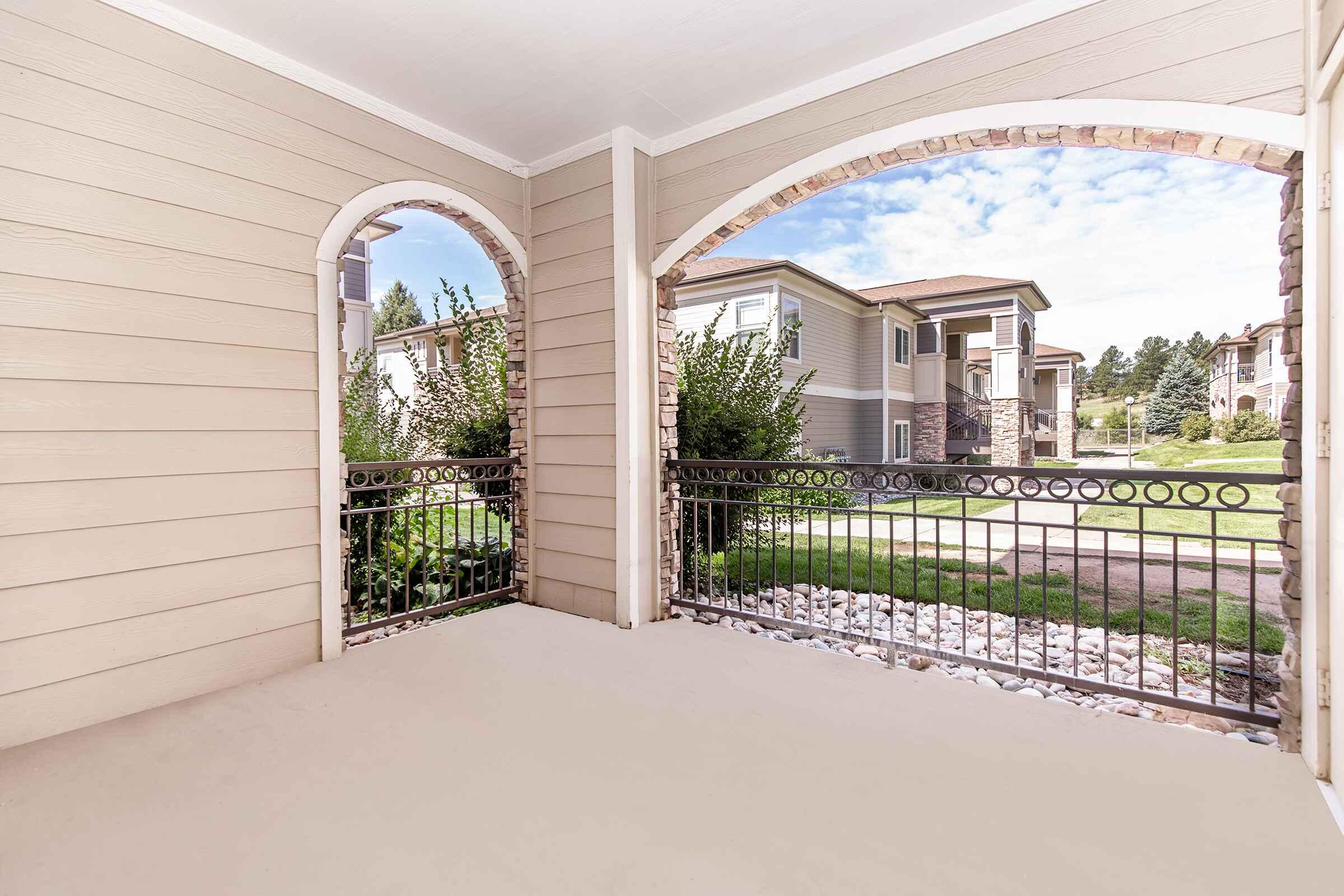 A view from a covered porch featuring two arched openings, with a railing made of wrought iron. The porch has a light-colored floor and opens up to a landscaped area with grass and stone pathways. In the background, there are residential buildings surrounded by greenery under a partly cloudy sky.