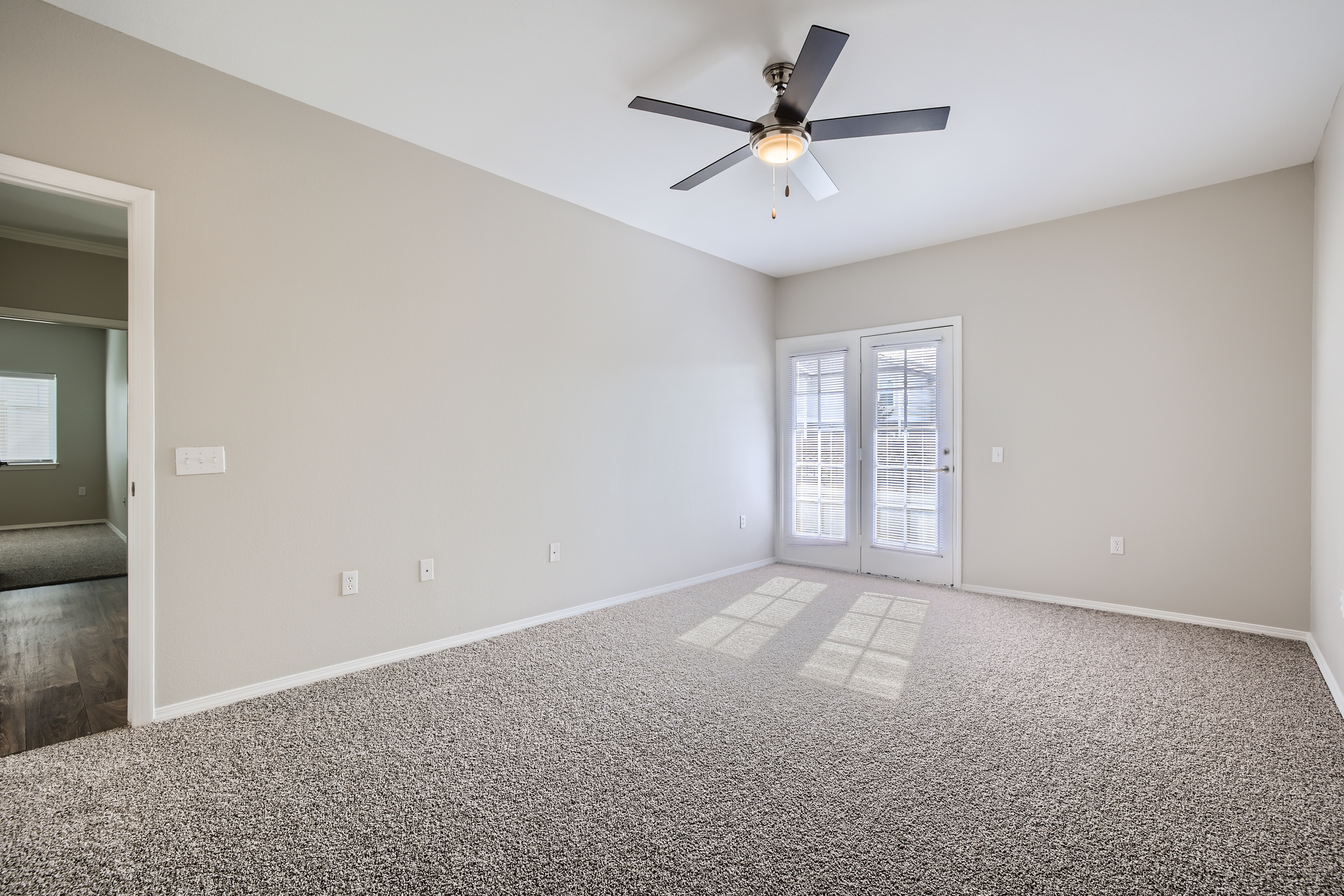 A spacious, empty room featuring beige walls and carpeted flooring. A ceiling fan is installed in the center, and there are double French doors leading to an outdoor area, allowing natural light to fill the space. Windows are visible in the background of another room.