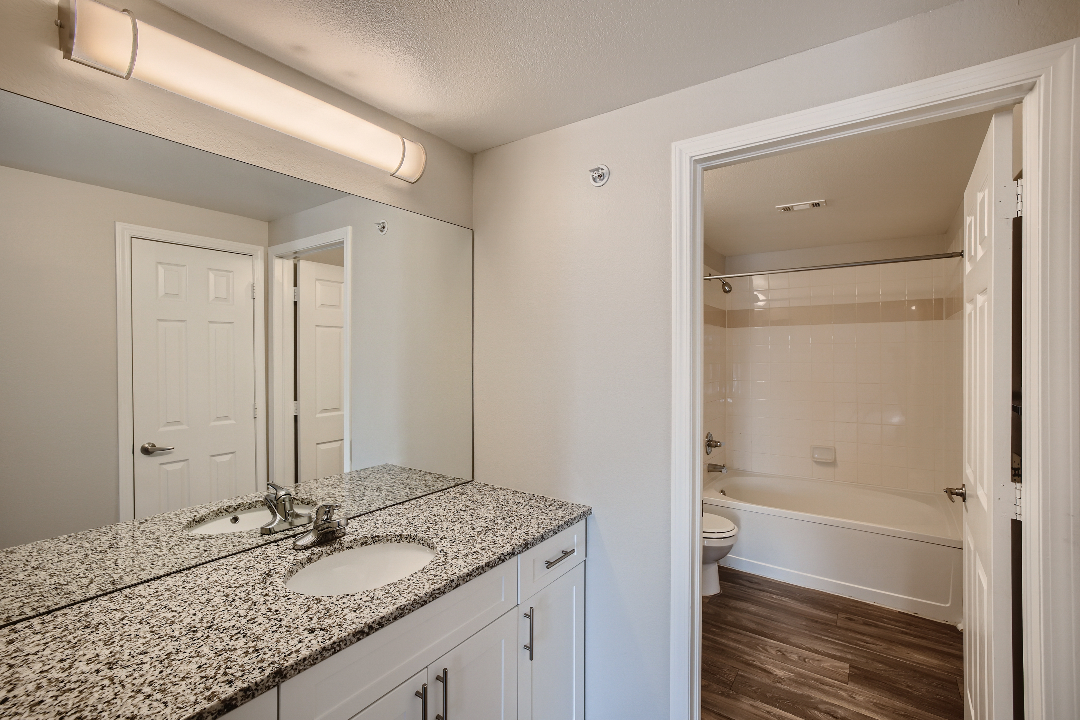 A well-lit bathroom featuring a large mirror above a granite countertop sink, white cabinetry, and a shower-tub combo visible through an open door. The flooring is dark wood, and there are white doors leading to other areas, enhancing the clean and modern aesthetic.