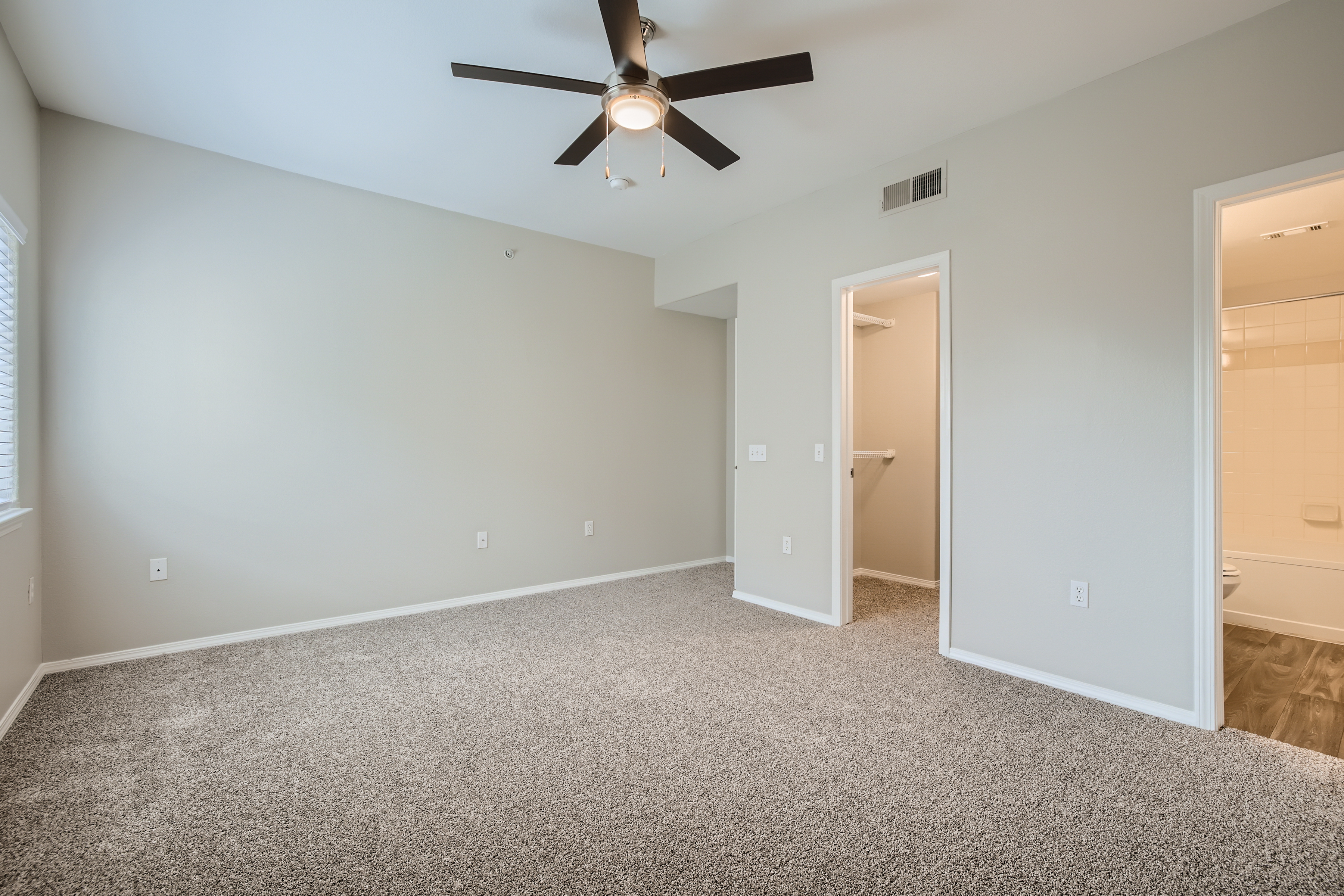 A spacious, empty room featuring light gray walls and carpeted flooring. A ceiling fan hangs from the ceiling, and there are two doorways: one leading to a bathroom and another potentially leading to a closet. Natural light comes in through a window on the left.