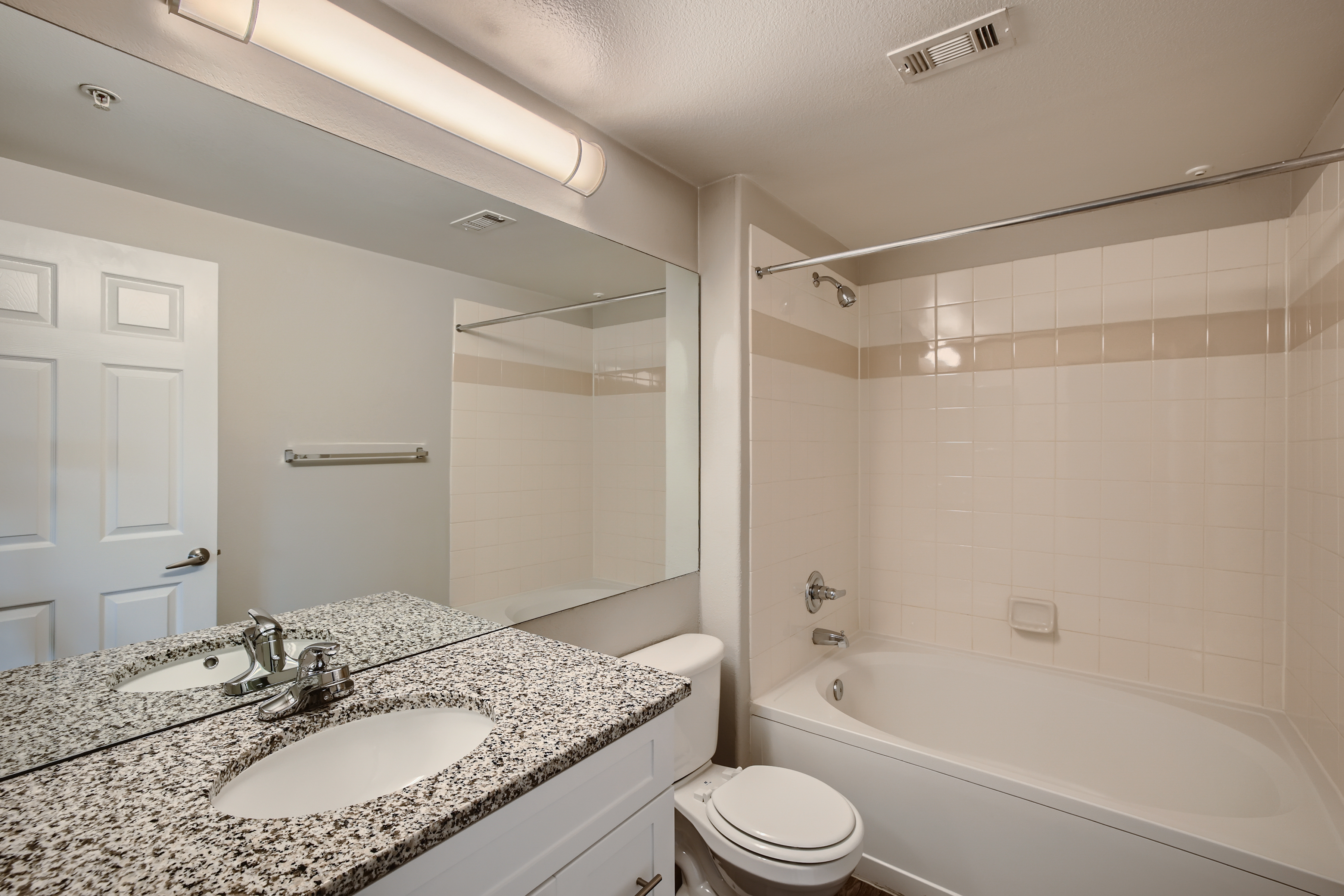 A clean, modern bathroom featuring a white tub with a shower curtain, a large mirror above a granite countertop sink, and a white toilet. The walls are tiled in light colors with a subtle accent stripe. There's a light fixture on the ceiling, and a towel rack is mounted beside the door.