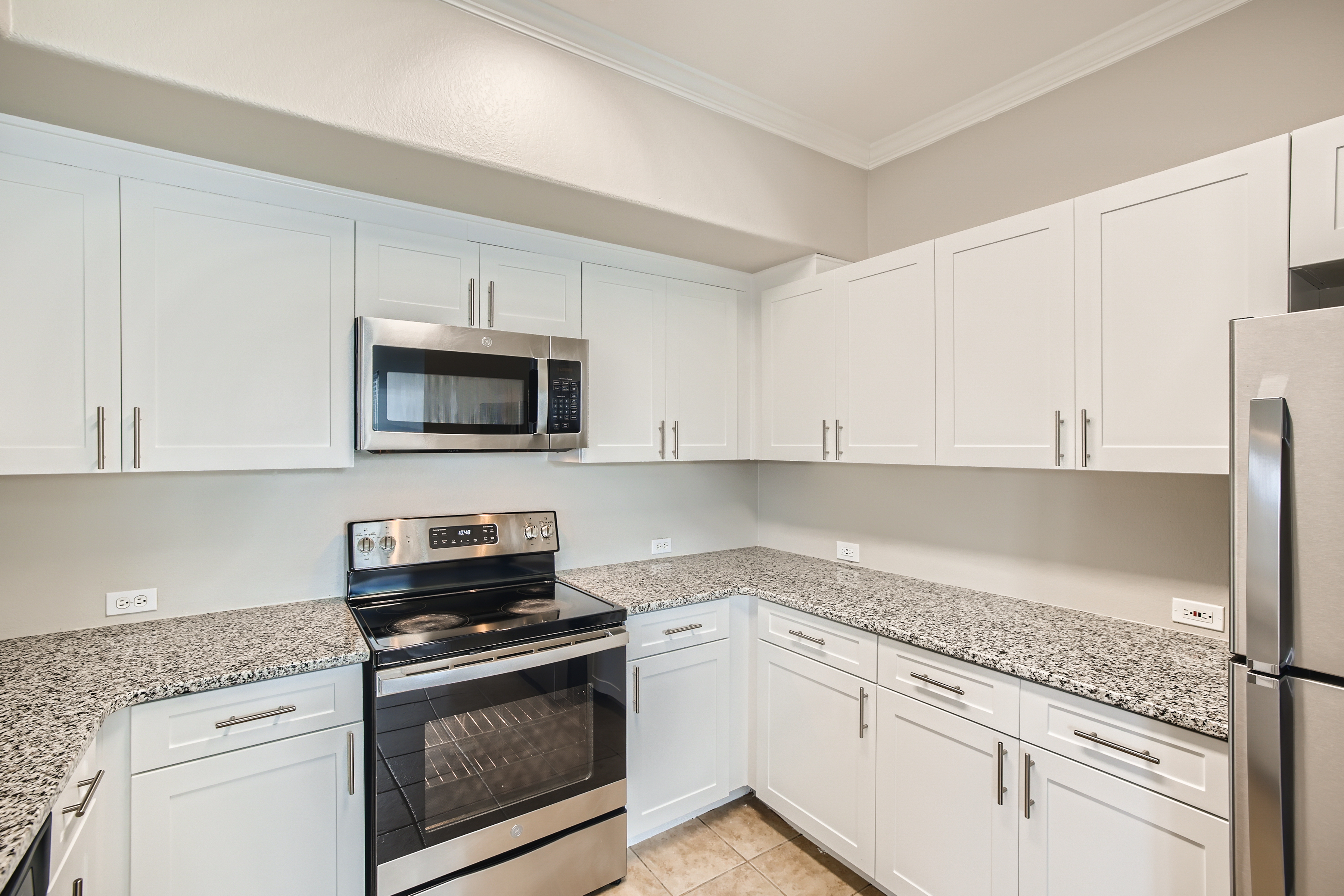 Modern kitchen featuring white cabinetry, a granite countertop, a stainless steel oven and microwave, and a side-by-side refrigerator. The space is well-lit with neutral wall colors and tiled flooring, providing a clean and contemporary aesthetic.