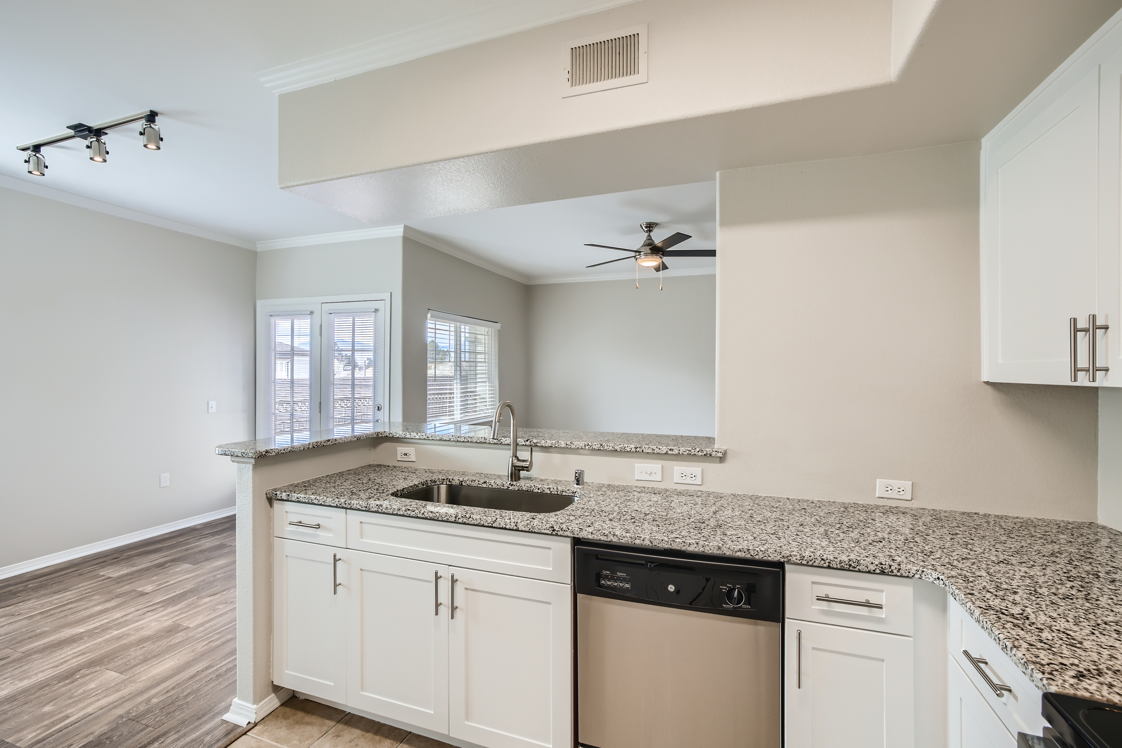 Modern kitchen featuring light gray walls, granite countertops, and white cabinetry. There is a stainless steel sink and a dishwasher. The space includes a ceiling fan and recessed lighting, with large windows allowing natural light to flood in. Wood-like flooring complements the design.