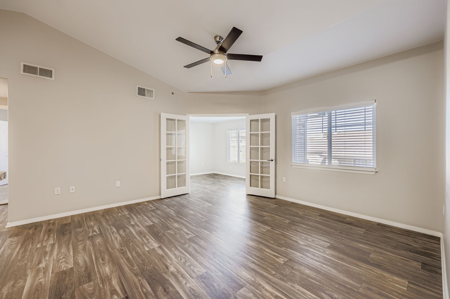 Spacious interior of a room with a ceiling fan, large windows, and wood-like flooring. There are light-colored walls and double French doors leading to another area. The overall ambiance is bright and modern, suitable for a living or entertainment space.