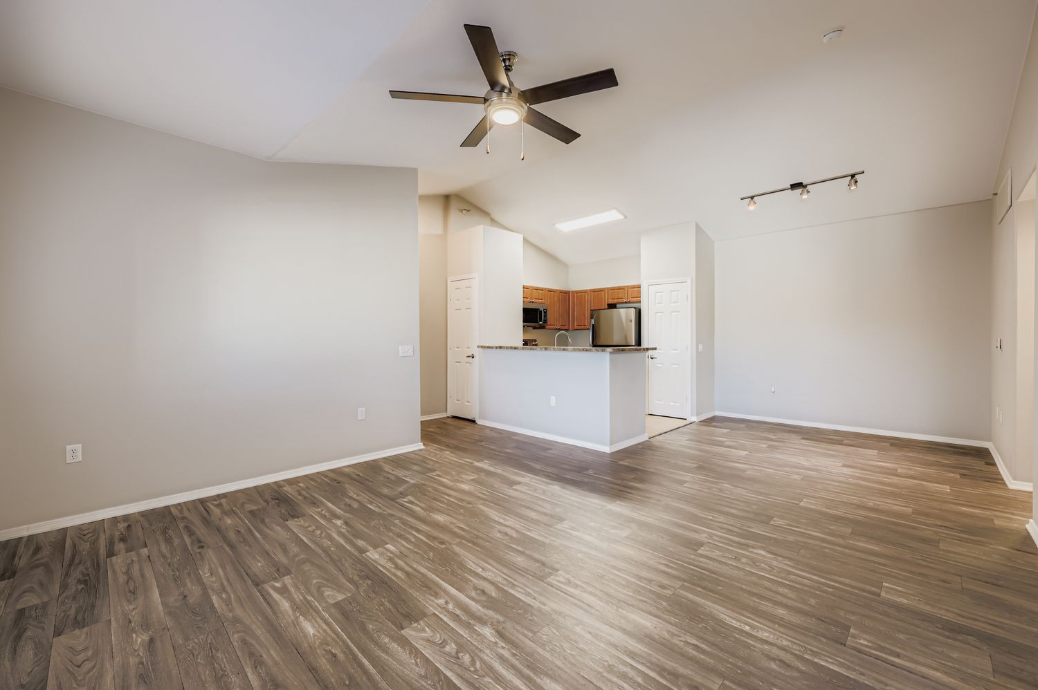 A spacious, modern living area with light wood-style flooring, featuring a ceiling fan. The open kitchen is partially visible with wooden cabinets and a stainless steel refrigerator. Bright, neutral-colored walls enhance the airy atmosphere, and there are recessed lighting fixtures on the ceiling.