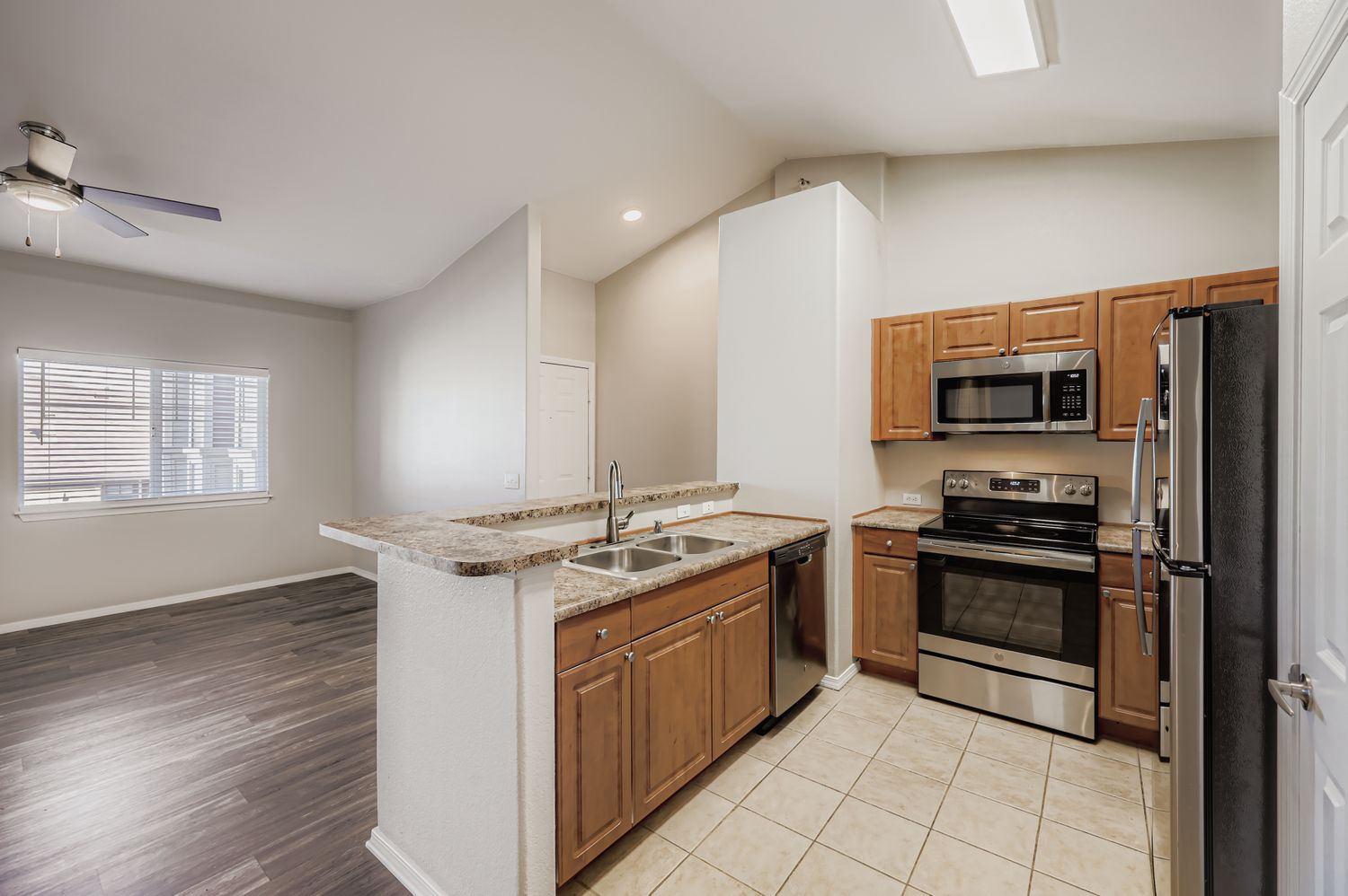 Modern kitchen showcasing wooden cabinetry, stainless steel appliances including a microwave and stove, and a double sink. The open layout features a view into a spacious living area with large windows and hardwood flooring, complemented by neutral-colored walls and high ceilings.