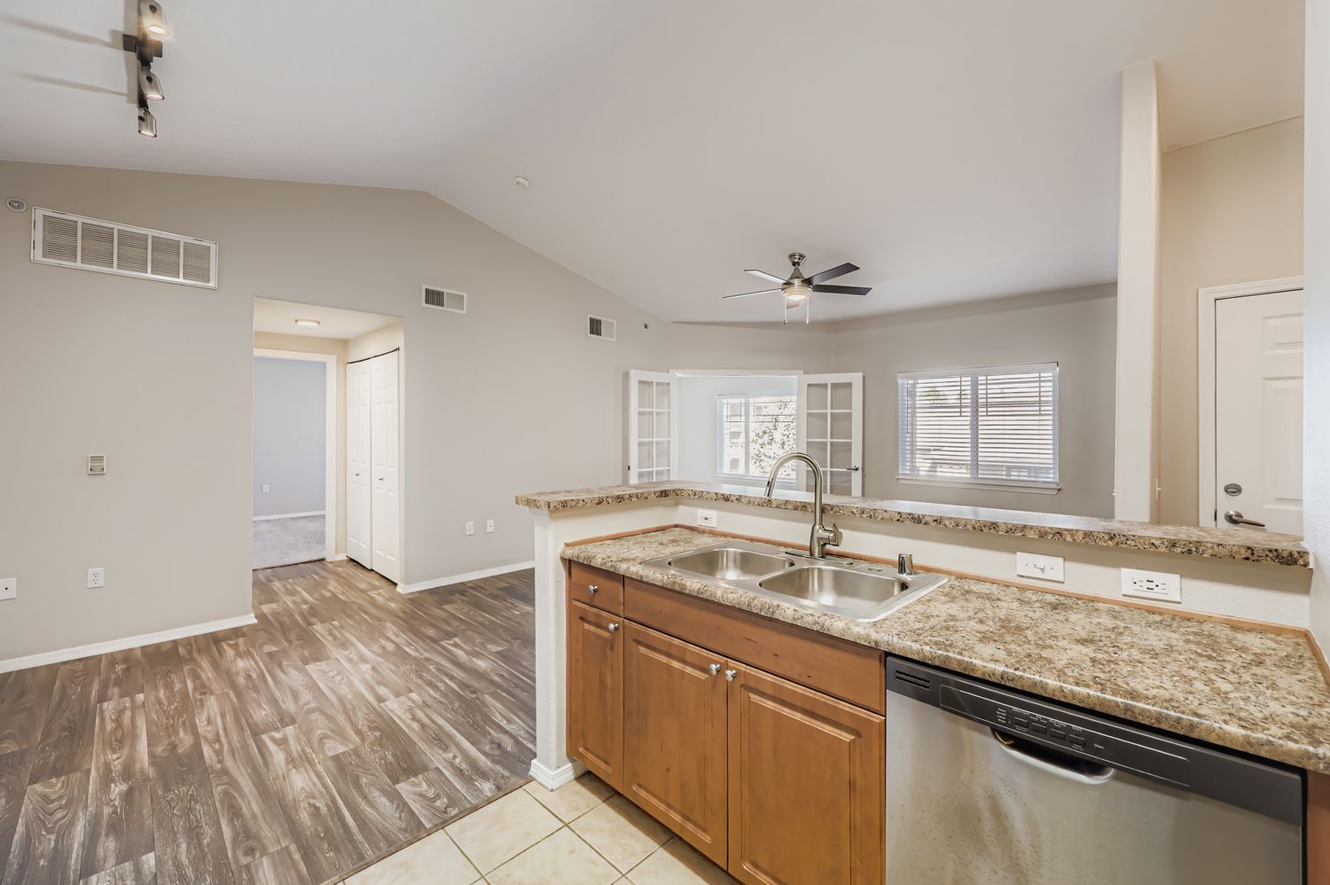 Open-concept kitchen showing wood cabinets, granite countertops, and stainless steel sink. A dishwasher is visible, and the space leads to a living area with a ceiling fan and large windows. The adjacent room has a door and closet, featuring neutral-colored walls and flooring.