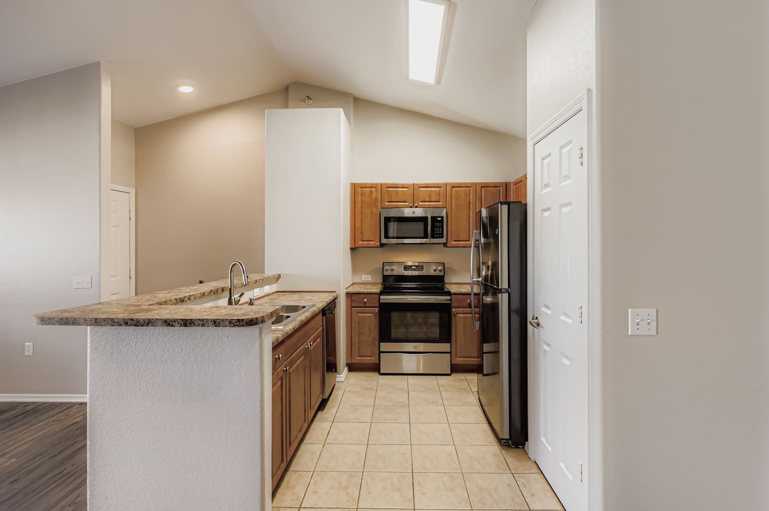 Modern kitchen with granite countertops, wooden cabinetry, and stainless steel appliances. The layout includes a stove, microwave, refrigerator, and an island with a sink. The walls are painted in neutral tones, and the room features high ceilings with a light fixture.