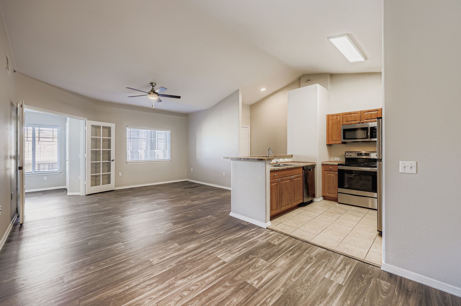 Spacious, modern living area with high ceilings, featuring a combined kitchen and dining space. The kitchen has wooden cabinetry, stainless steel appliances, and a breakfast bar. Large windows provide natural light, and there's access to a room through double doors on the left. The flooring is a blend of laminate and tile.