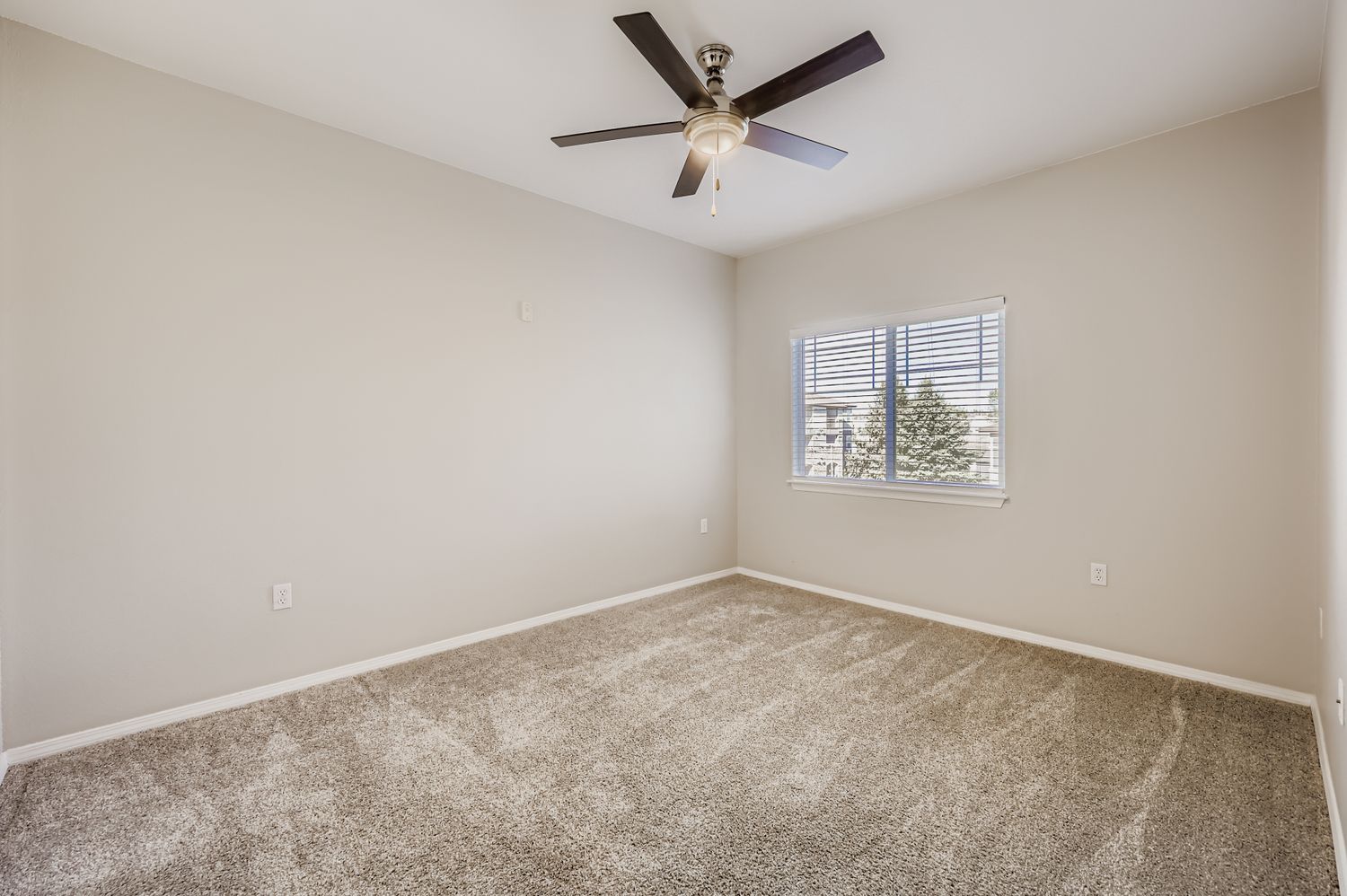 Empty room with light beige walls and a ceiling fan. The floor is carpeted in a similar beige tone. A window with blinds allows natural light to enter, providing a view of trees outside. The room appears spacious and ready for furnishing.