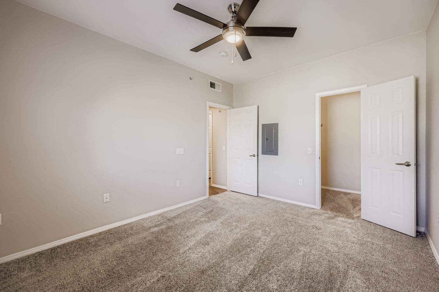 A spacious, well-lit bedroom featuring light gray walls, plush carpet, and a ceiling fan. Two white doors are visible, leading to closets or other rooms, with a modern light switch on the wall. The room has a bright, airy feel, ideal for a cozy living space.