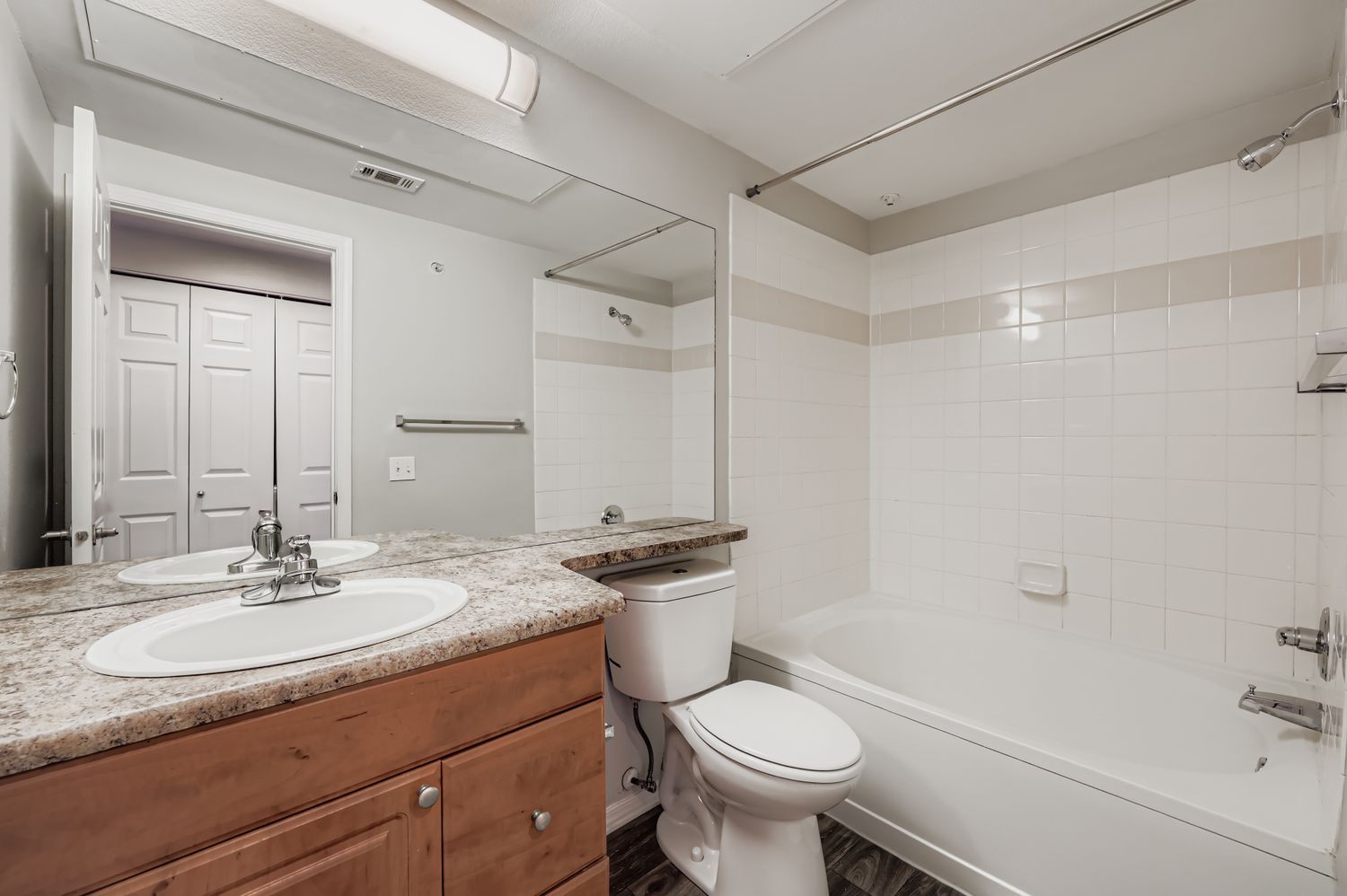 A modern bathroom featuring a double sink vanity with a granite countertop, a white toilet, and a bathtub with a shower. The walls are tiled with a light-colored backsplash, and there's a large mirror above the sinks. A door leads to a closet space, enhancing the overall functionality of the room.