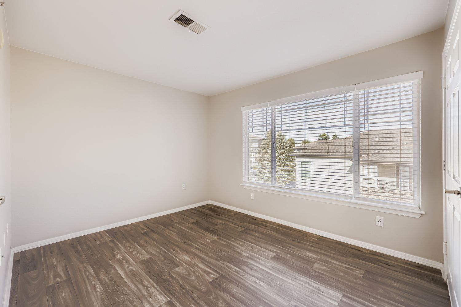 Empty room with light beige walls and laminate flooring. A large window with white blinds allows natural light, showing a view of the exterior. The space is unfurnished and clean, designed for potential use as a bedroom or office.